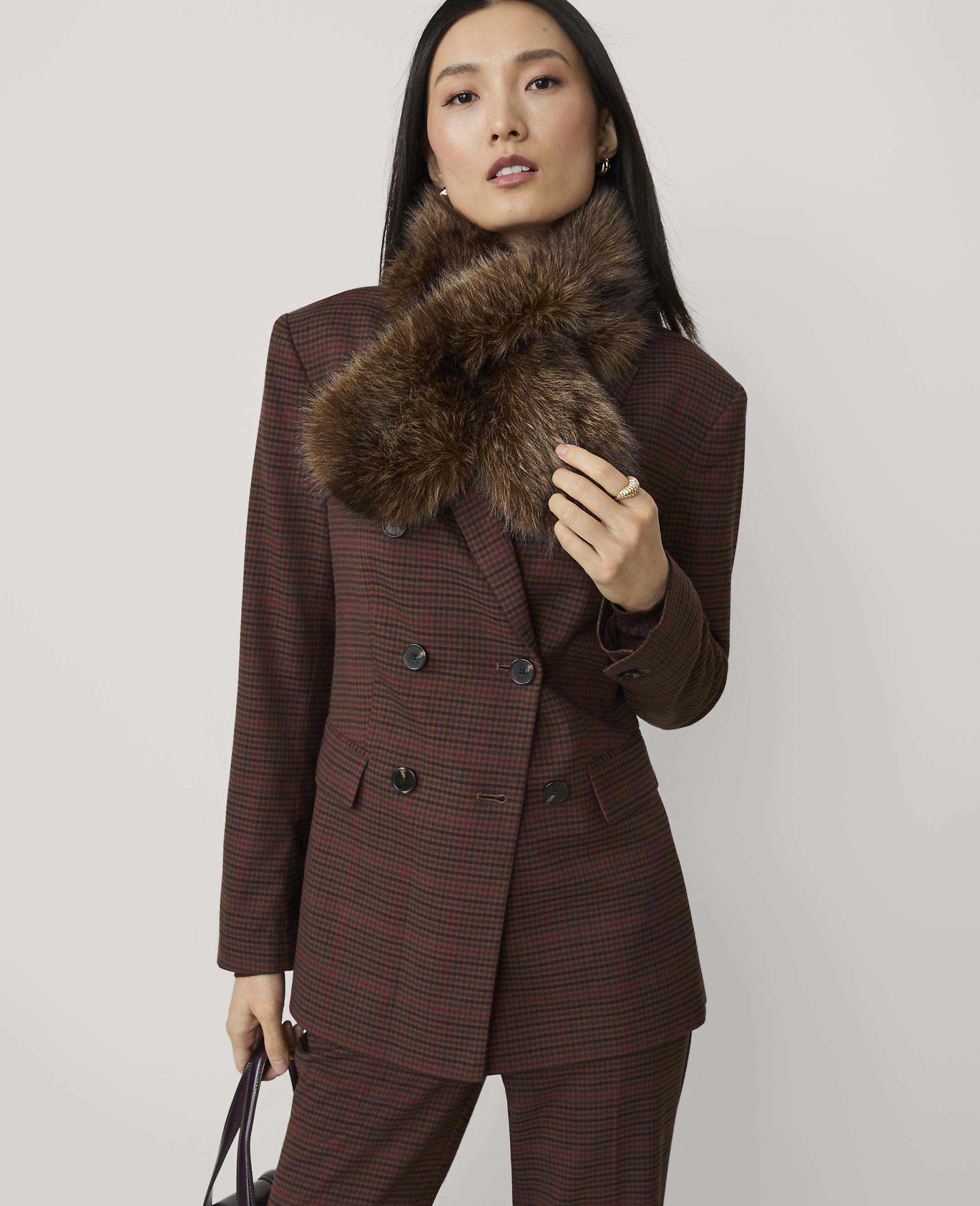 A woman in a brown plaid suit and fur collar stands against a plain gray background.