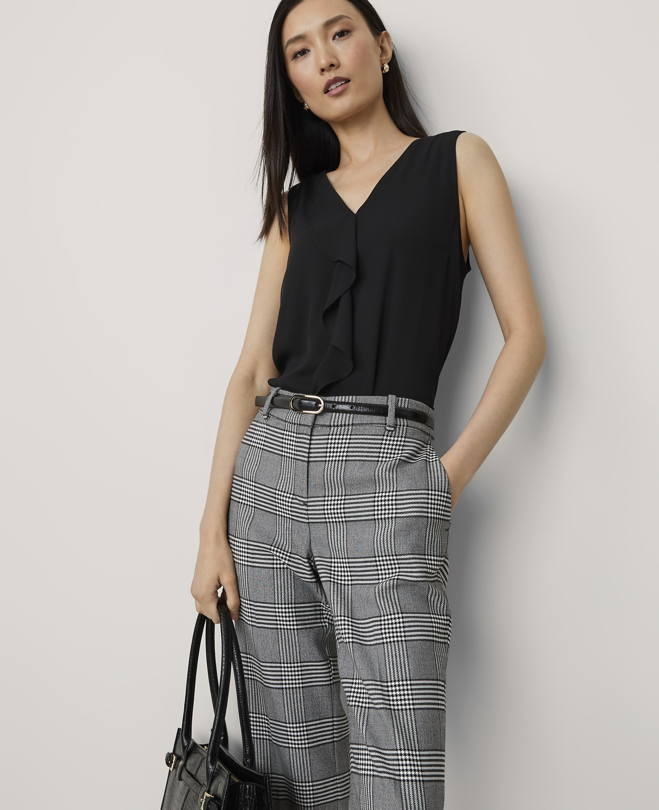 A woman in a black ruffled blouse and plaid pants stands against a plain white background, holding a black handbag.