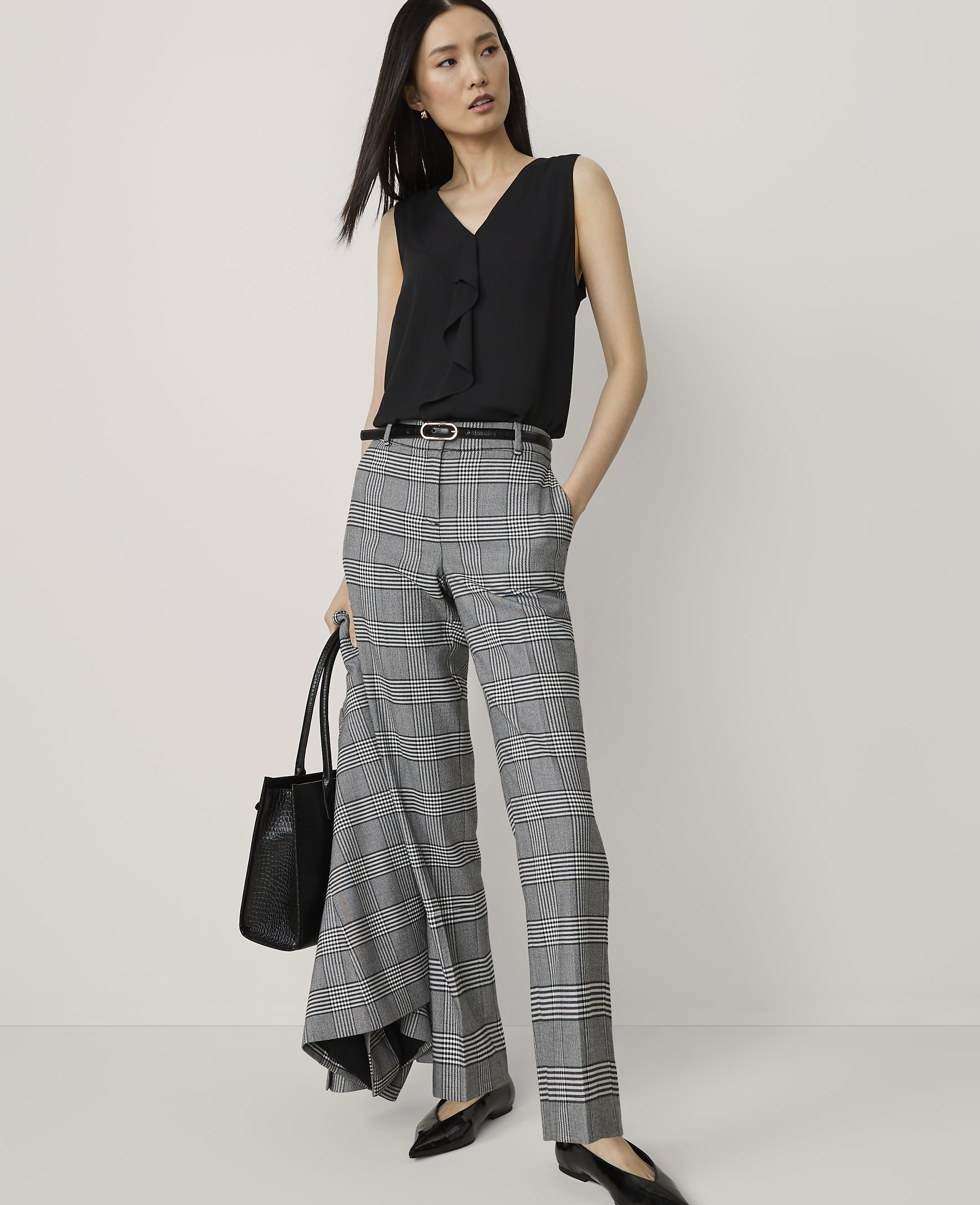 A woman in a black sleeveless top and plaid pants stands with a handbag in front of a plain white background.