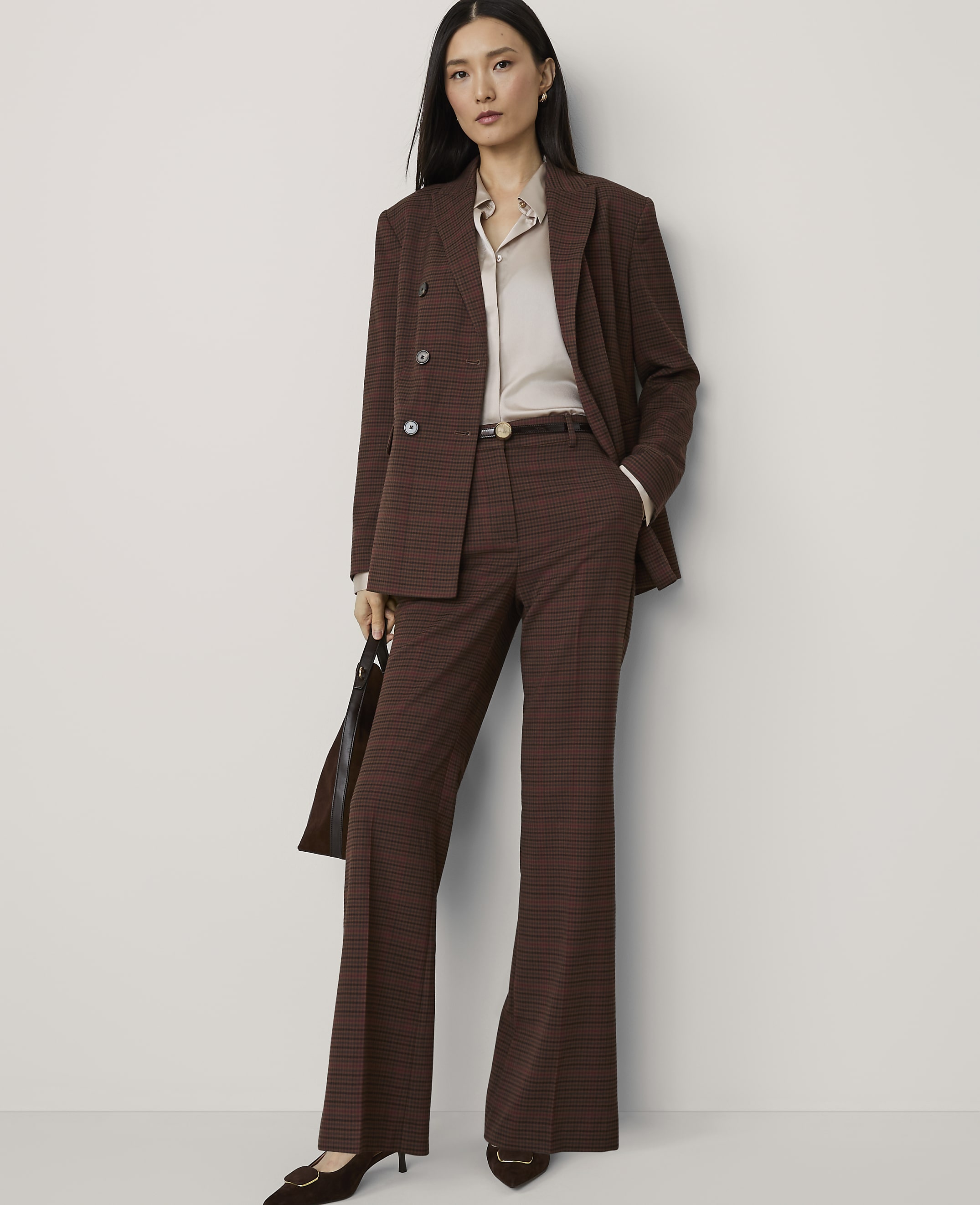 A woman in a brown plaid suit stands against a plain, light-colored background, holding a dark handbag.