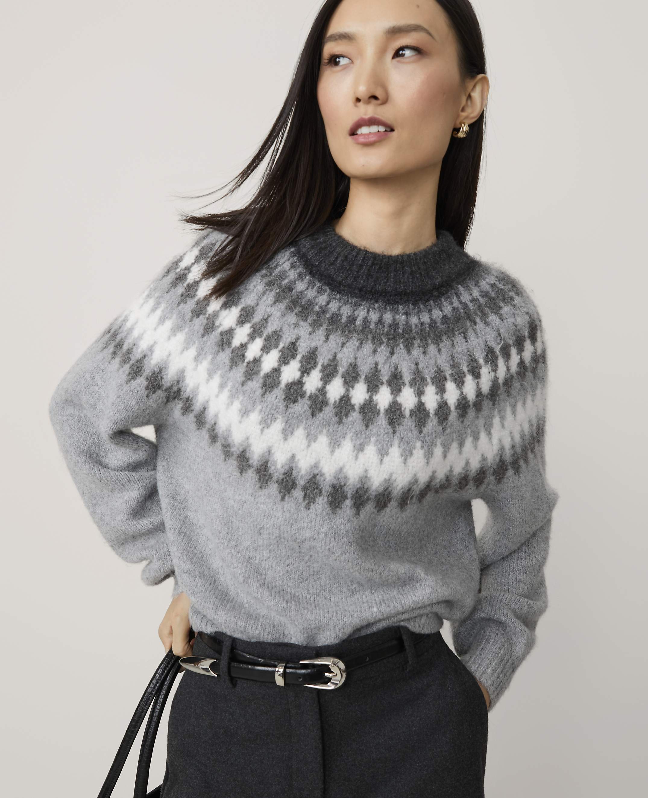 A woman in a grey fair isle sweater and black belt stands against a plain, light grey background.