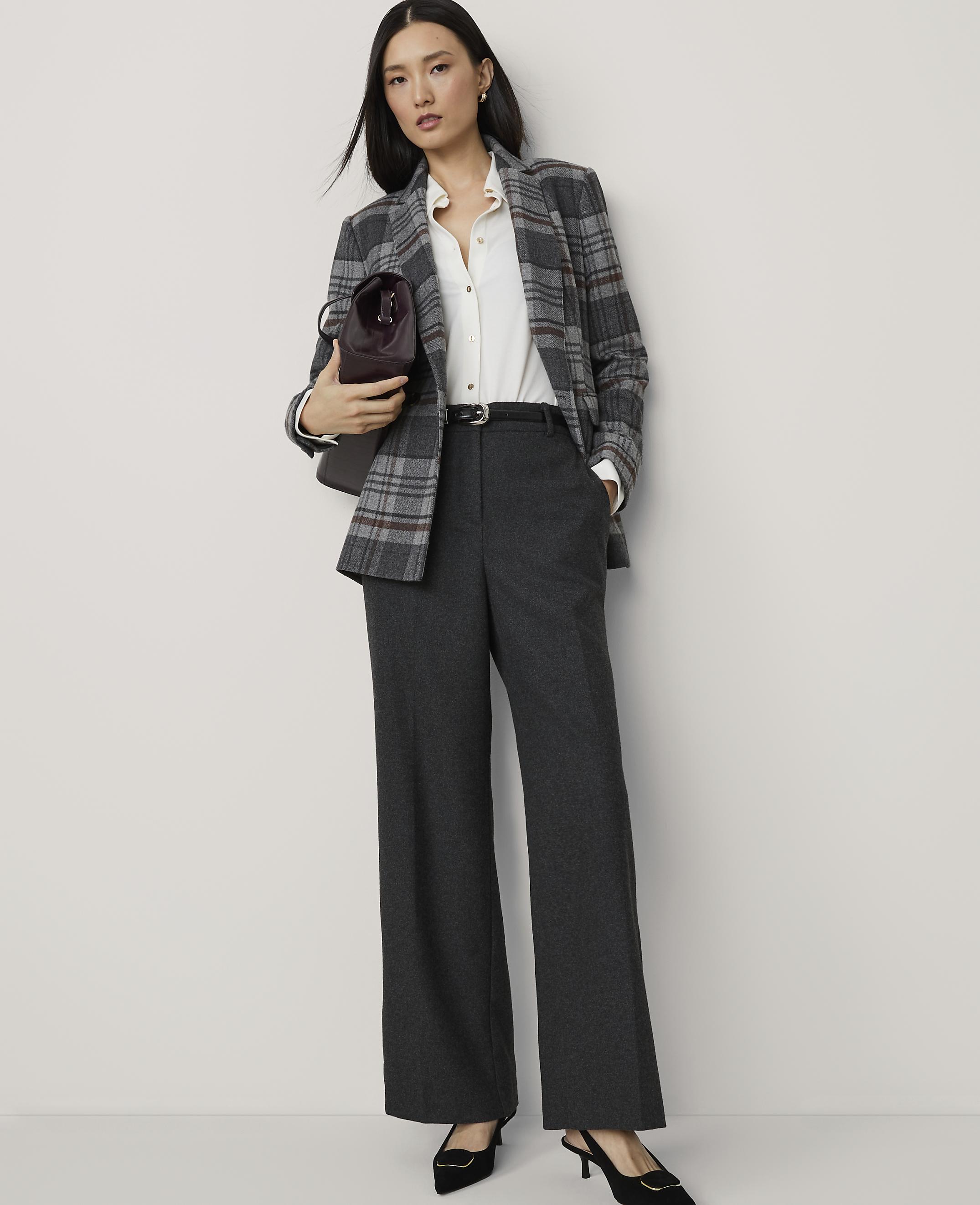 A woman in a plaid blazer and wide-leg pants stands against a plain white background, holding a burgundy handbag.