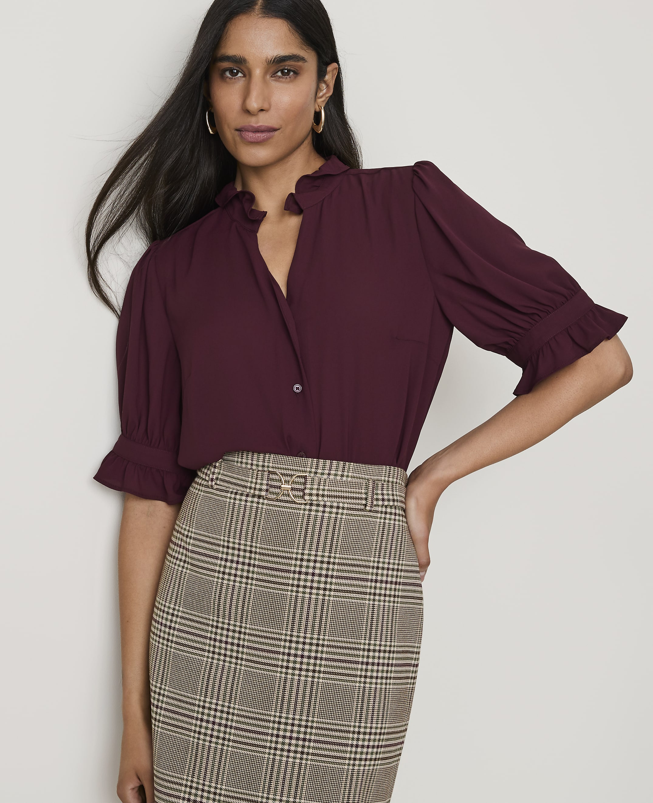 A woman in a burgundy ruffled blouse and plaid skirt stands against a plain white background.