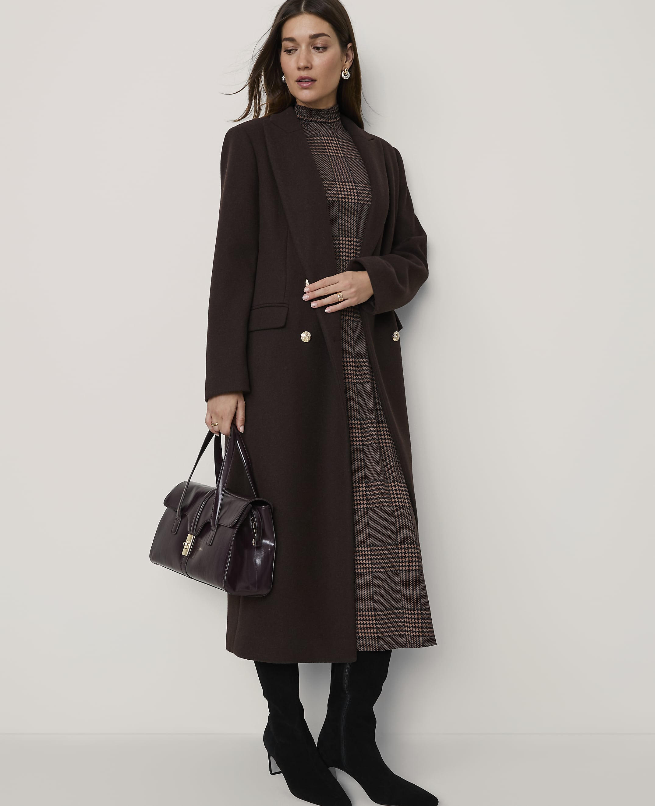 A woman in a brown coat and plaid dress stands against a plain wall, holding a dark handbag.