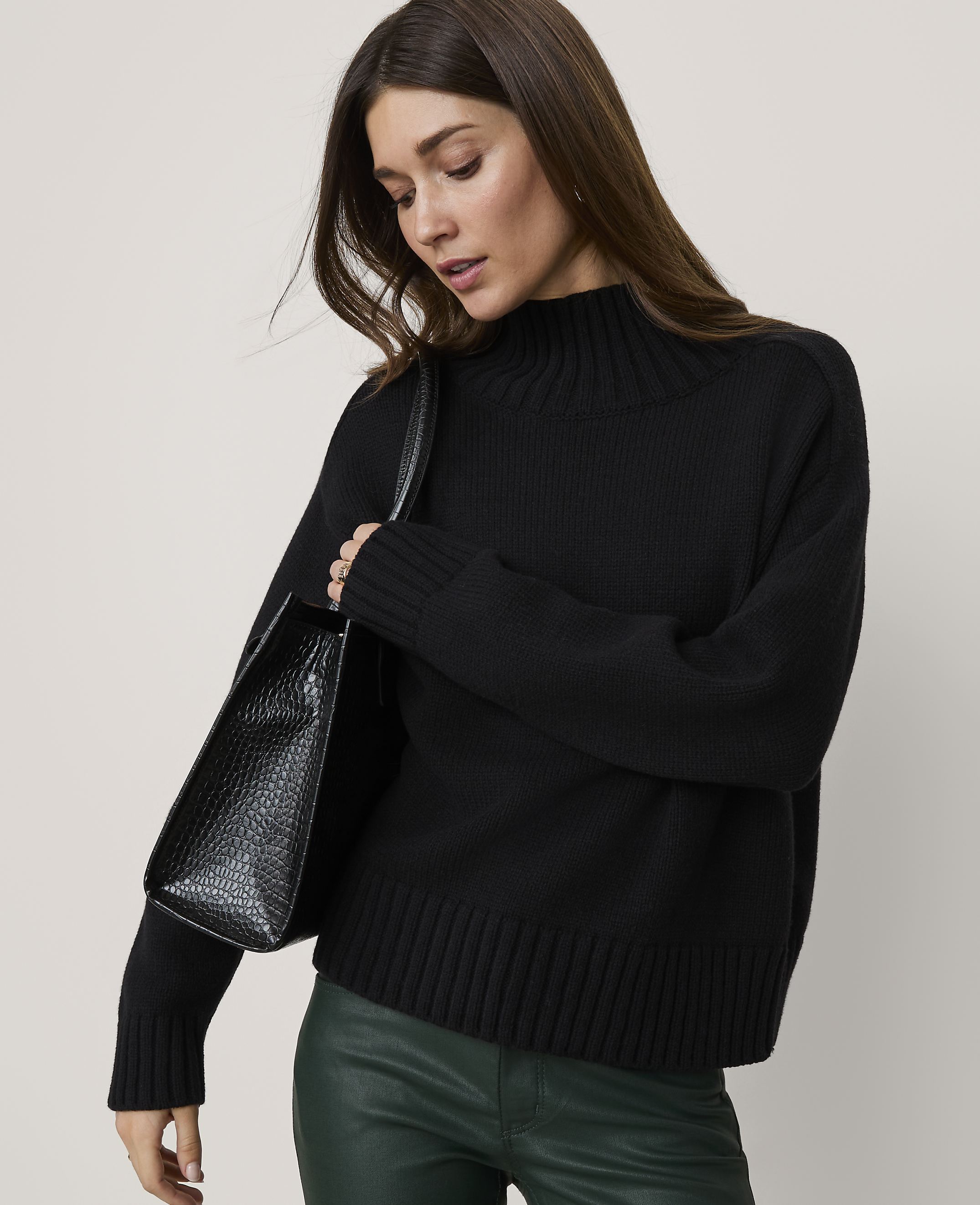 A woman in a black turtleneck sweater and green leather pants carries a black textured handbag against a plain gray background.