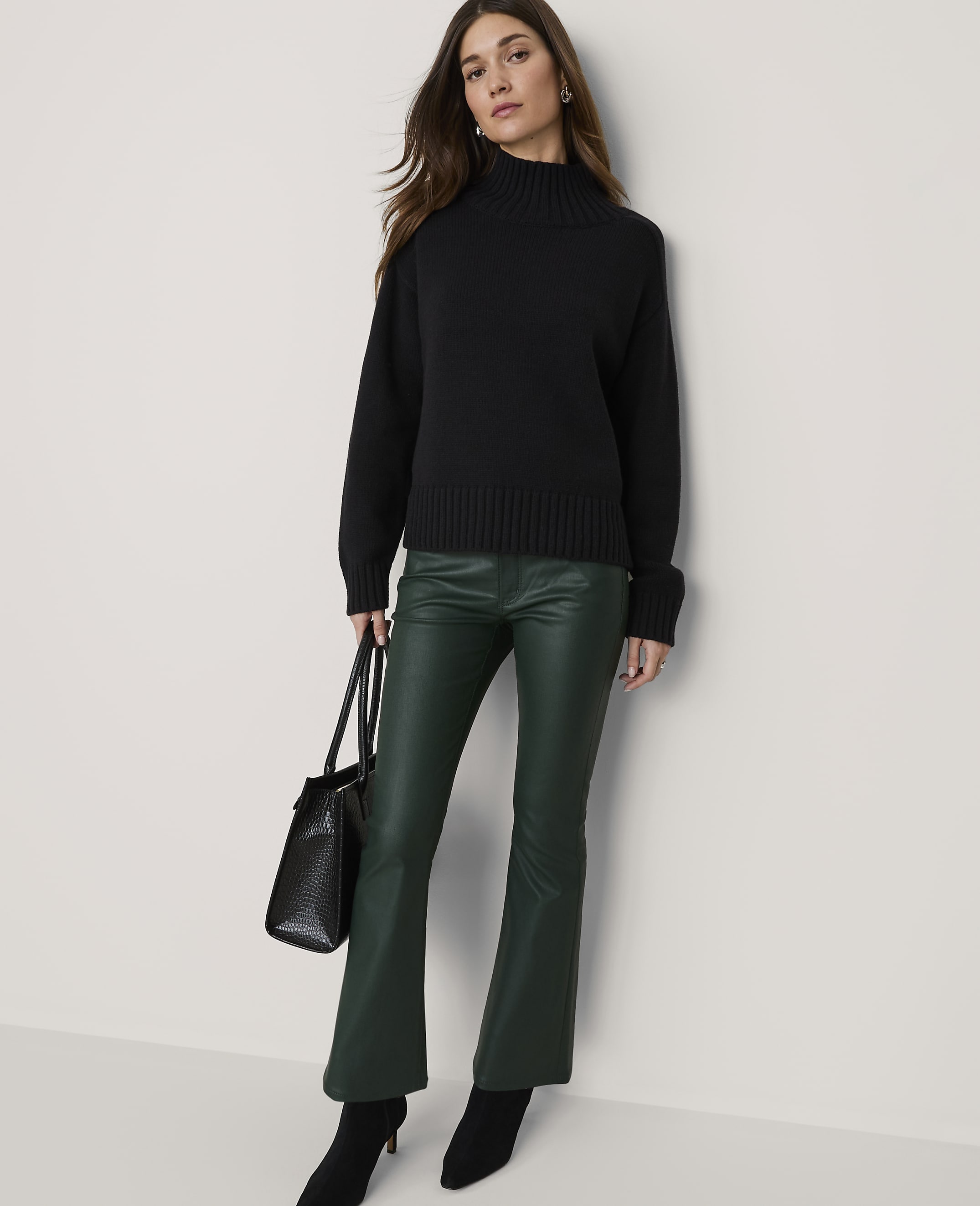 A woman in a black turtleneck and green flared pants stands against a plain, light-colored wall, holding a black handbag.