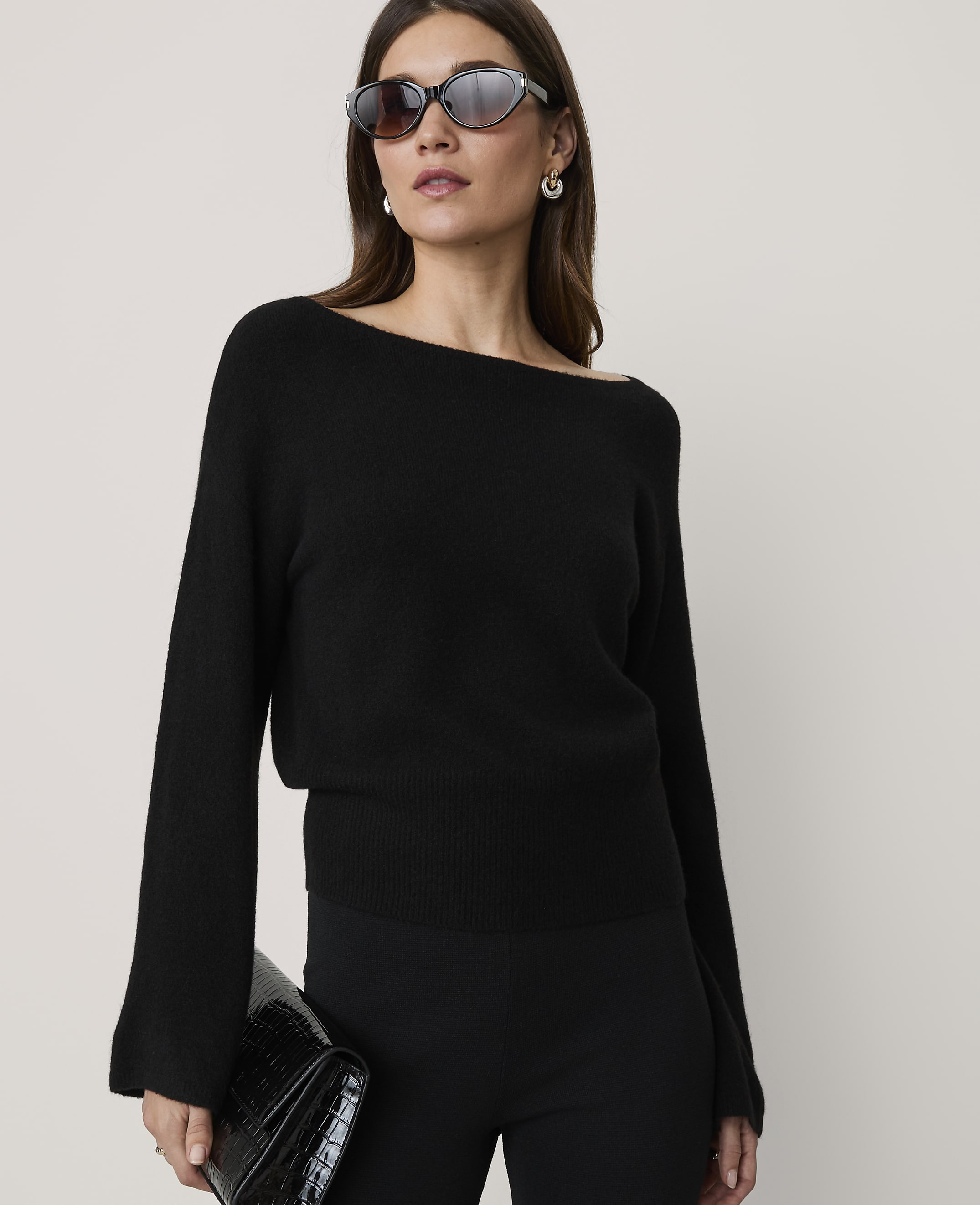 A woman in a black off-the-shoulder sweater and sunglasses holds a black clutch in front of a plain background.