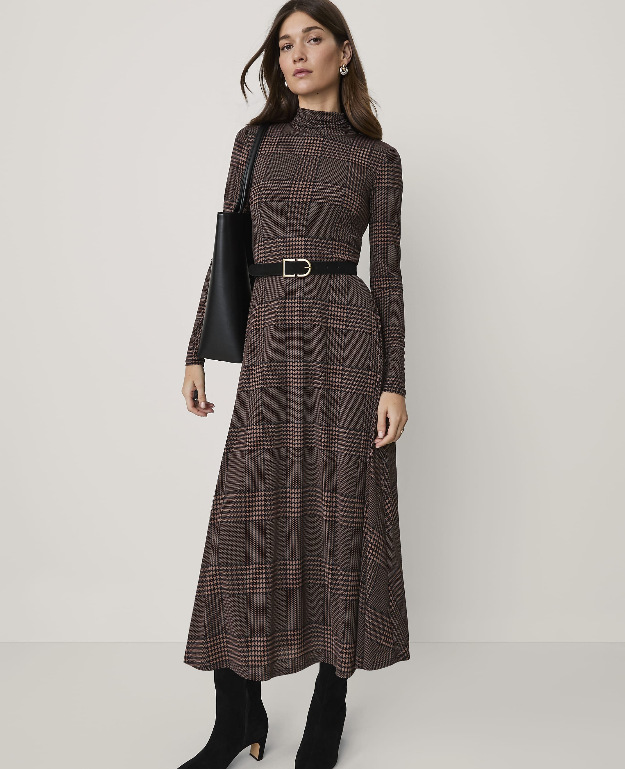 A woman in a brown plaid dress, black belt, and boots stands against a plain background.