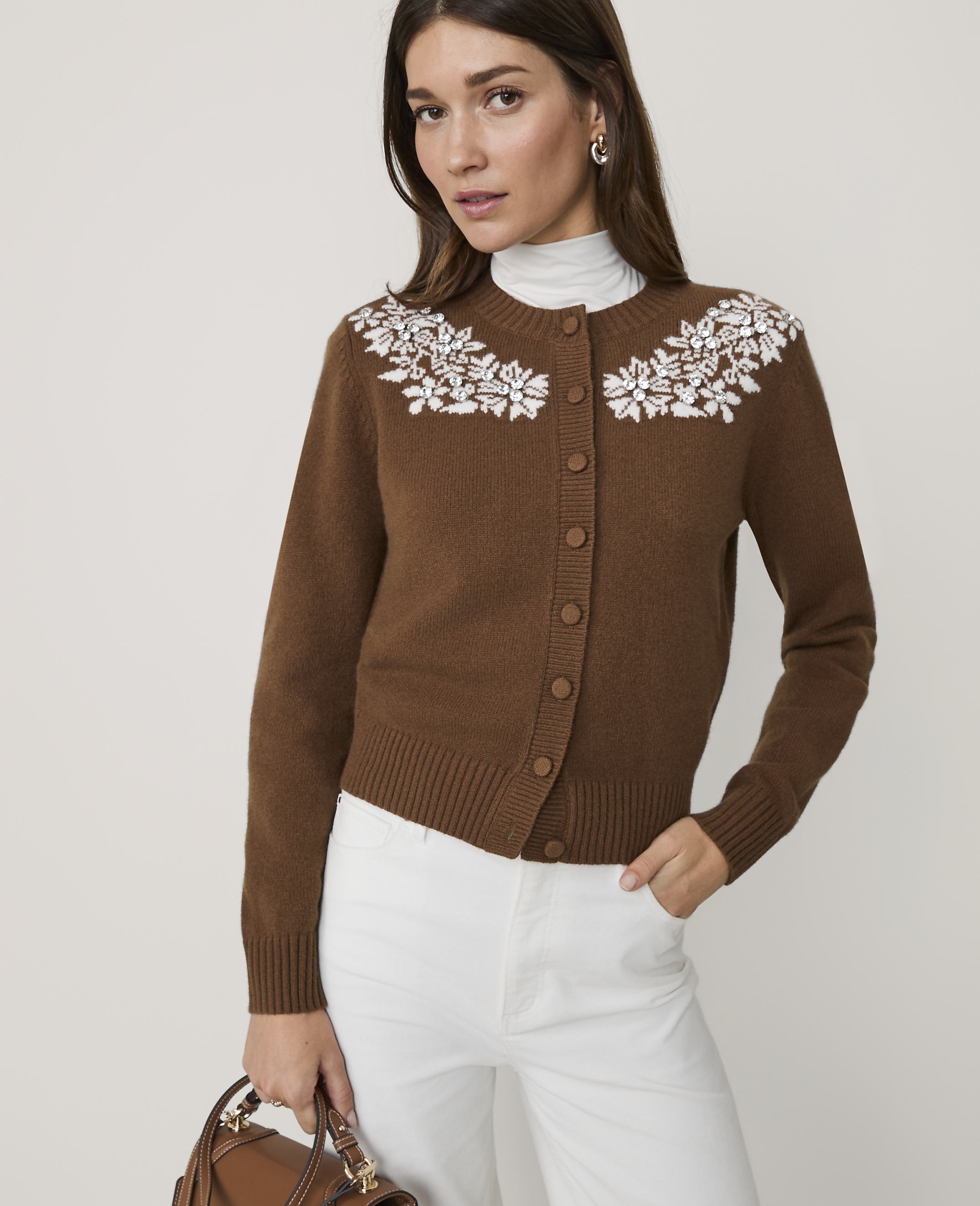 A woman in a brown cardigan with white floral embroidery and white pants stands against a plain white background, holding a brown handbag.