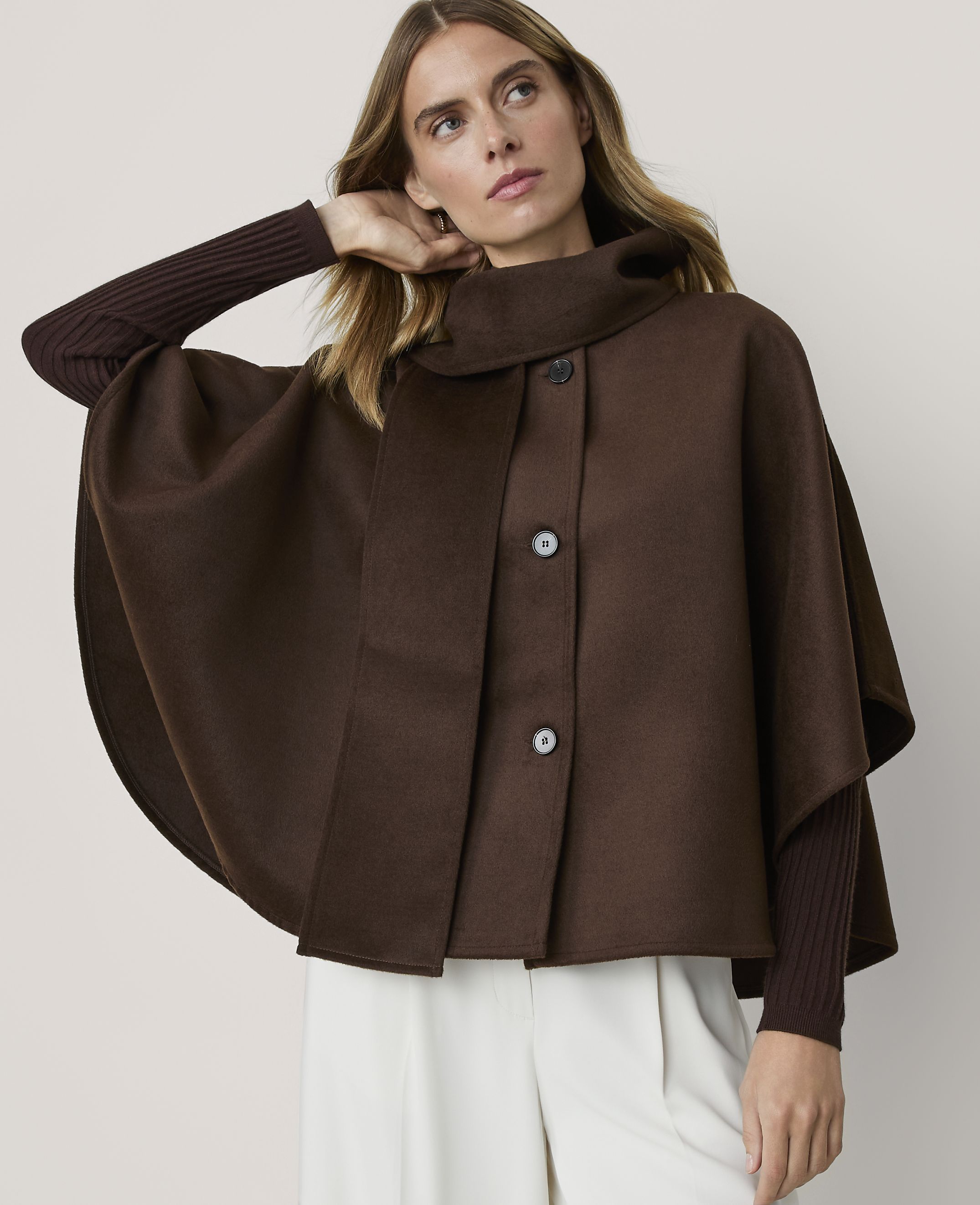 A woman in a brown cape and ribbed sleeves stands against a plain, light background.