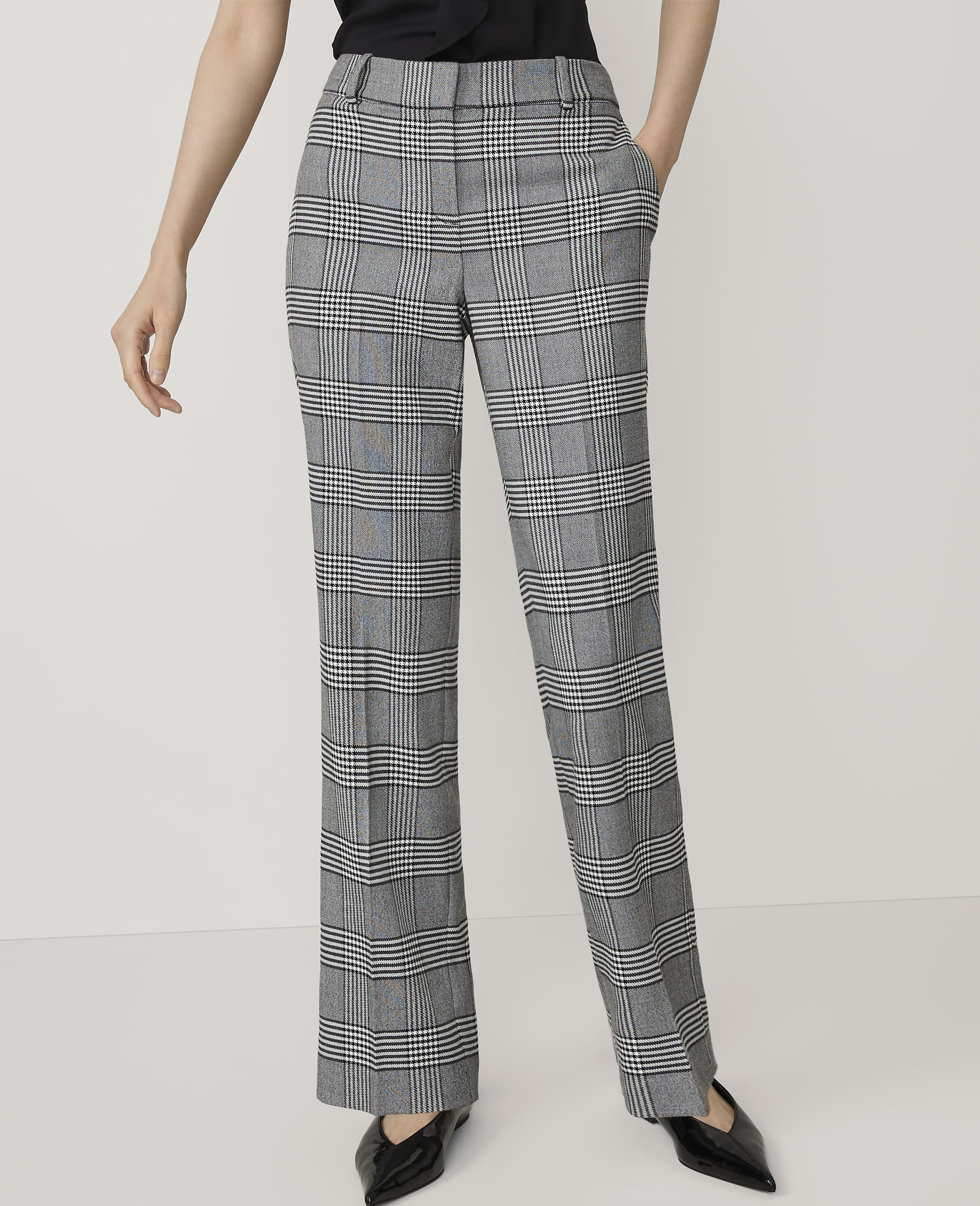 A person stands in the foreground wearing plaid trousers and black pointed-toe shoes against a plain white background.