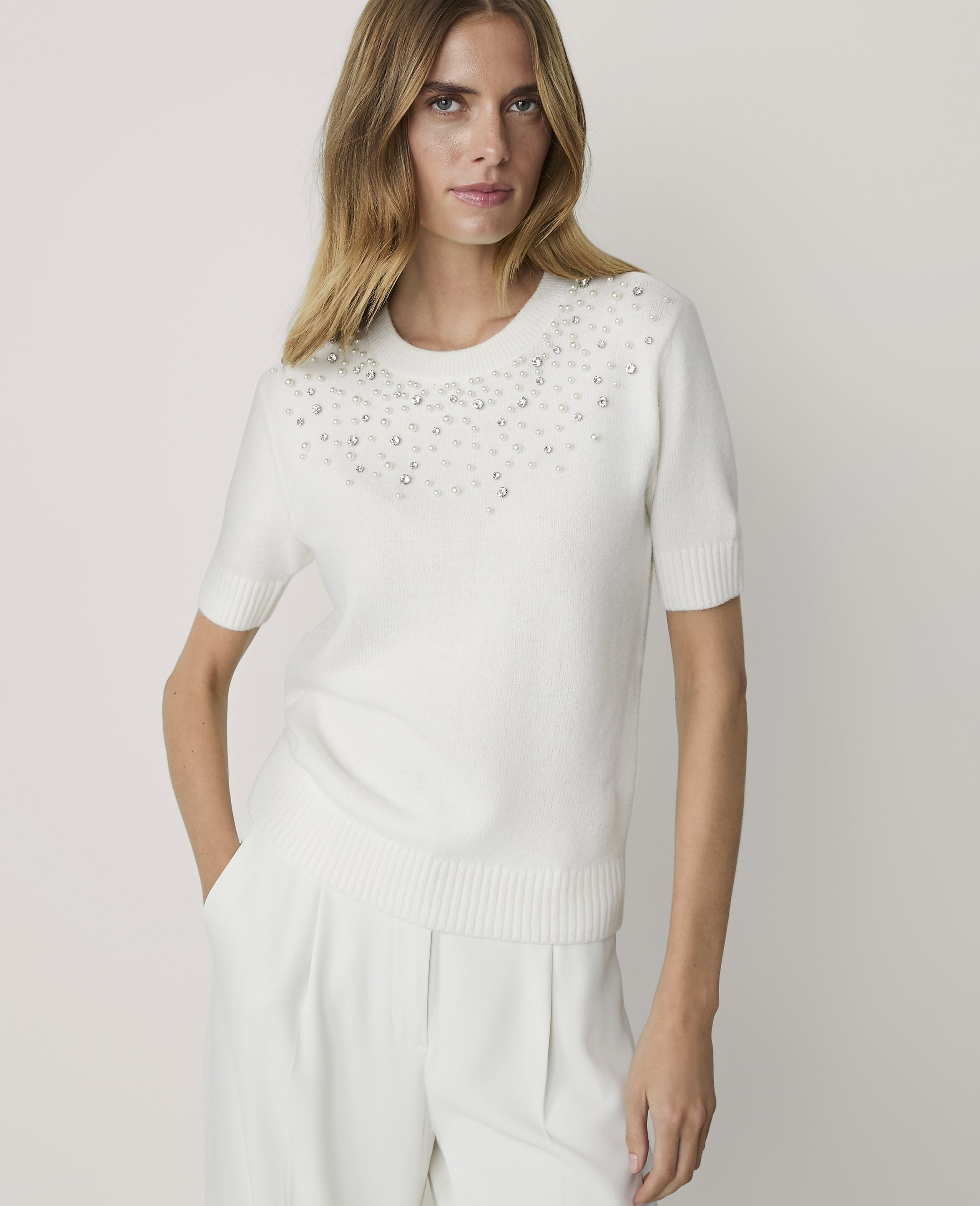 A woman in a white pearl-embellished sweater and white pants stands against a plain white background.