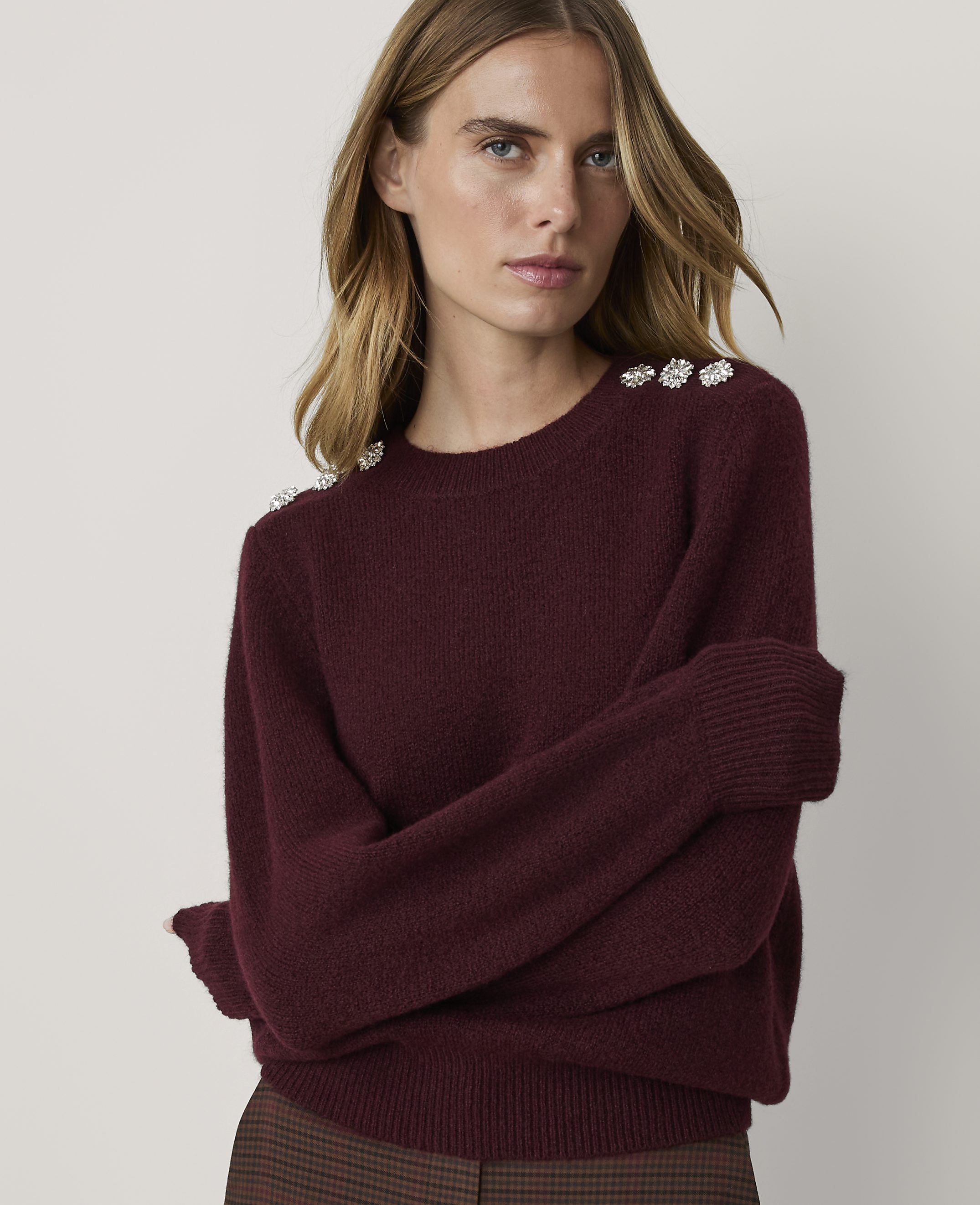 A woman in a maroon sweater with jeweled shoulders poses against a plain, light gray background.