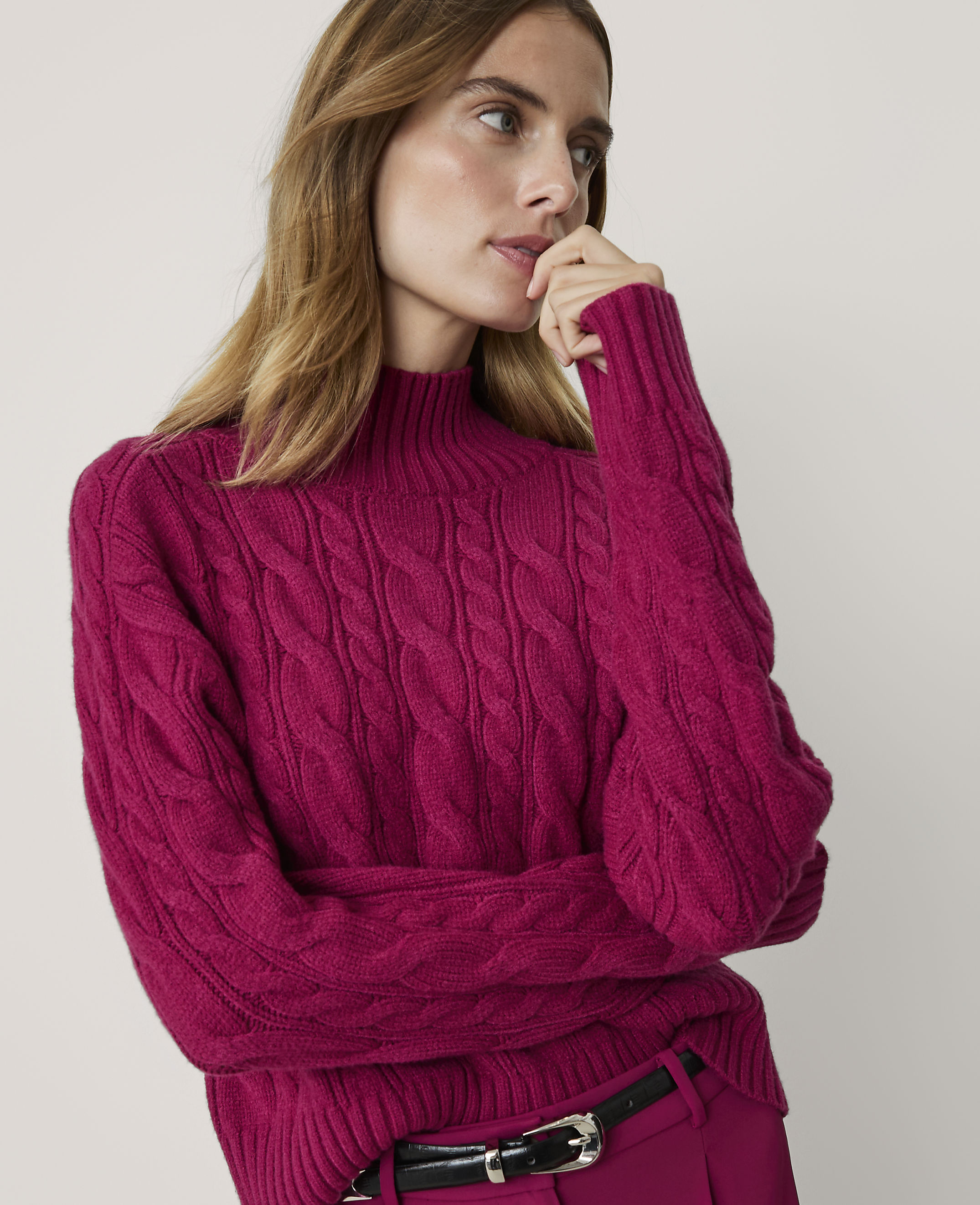 A woman in a magenta cable-knit sweater and pink pants poses against a plain white background.