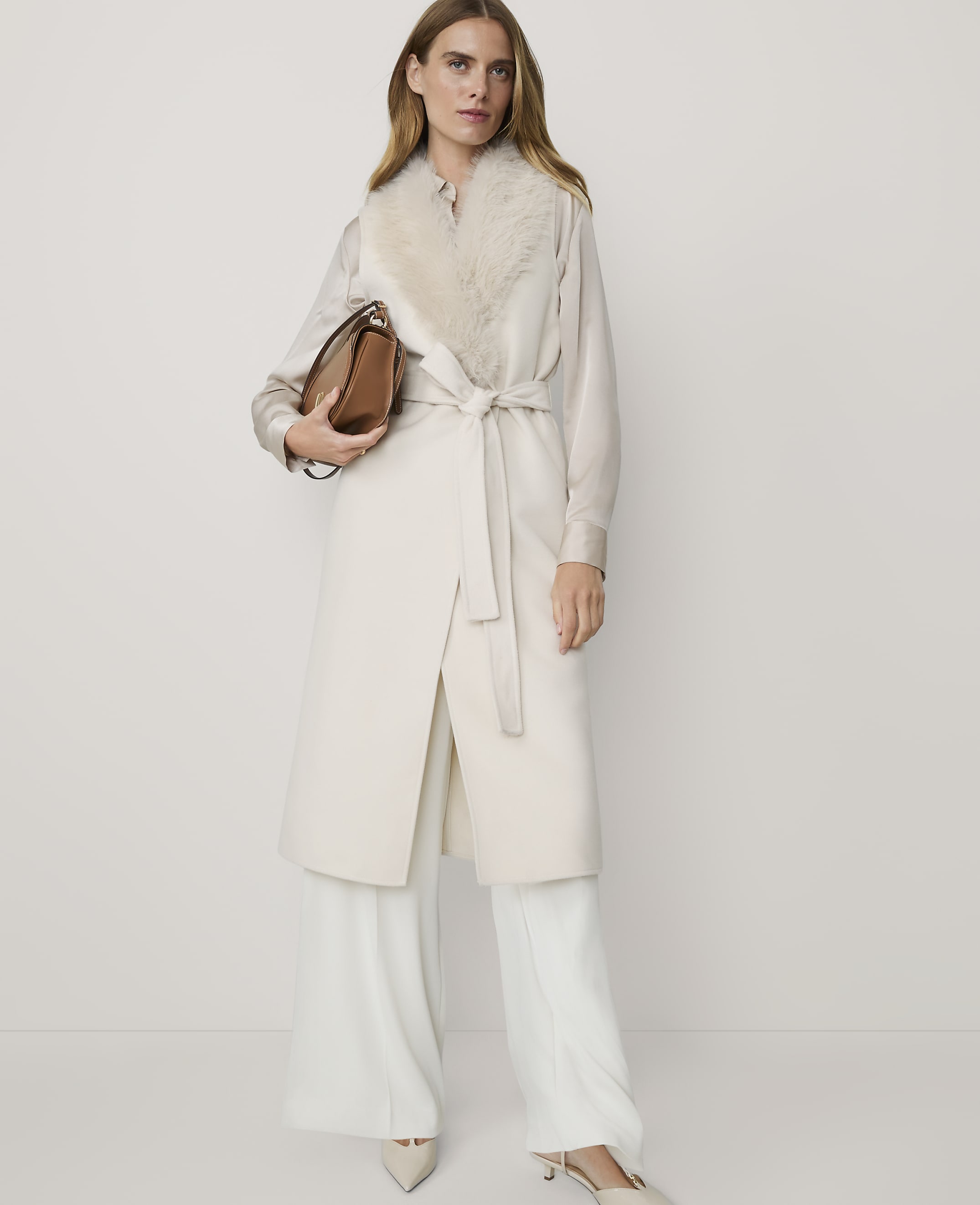 A woman in a cream coat with a fur collar and wide-leg pants stands against a plain, light background, holding a brown handbag.
