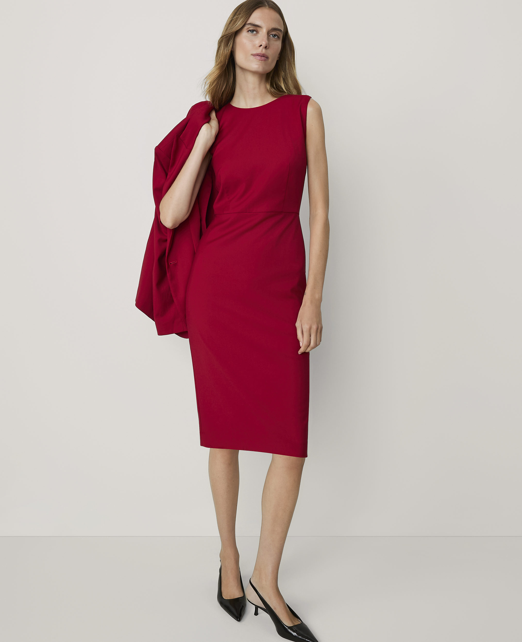 A woman in a red dress and black heels stands against a plain white background, with a red blazer draped over her shoulder.