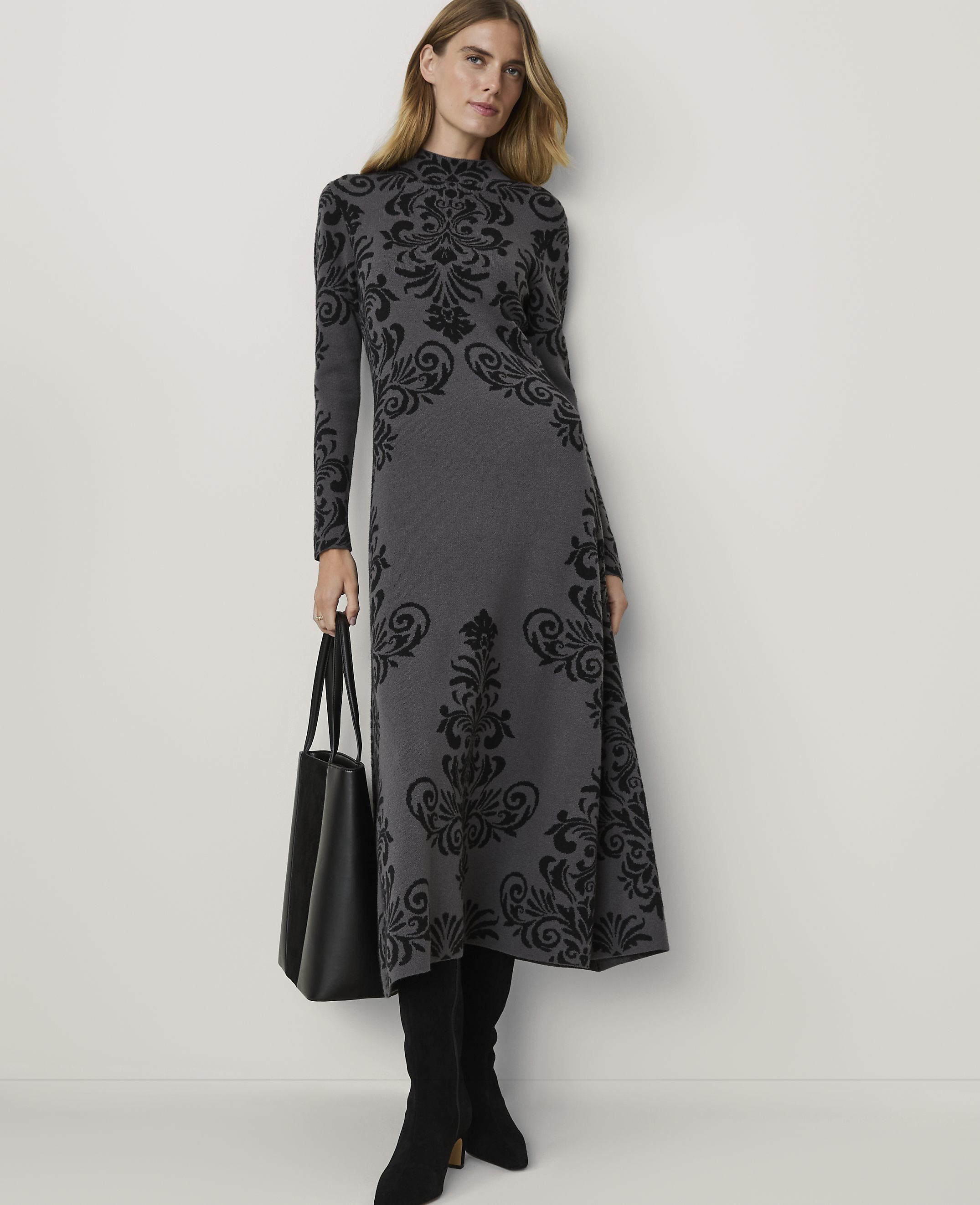 A woman in a grey patterned dress and black boots stands with a black tote bag against a plain white background.