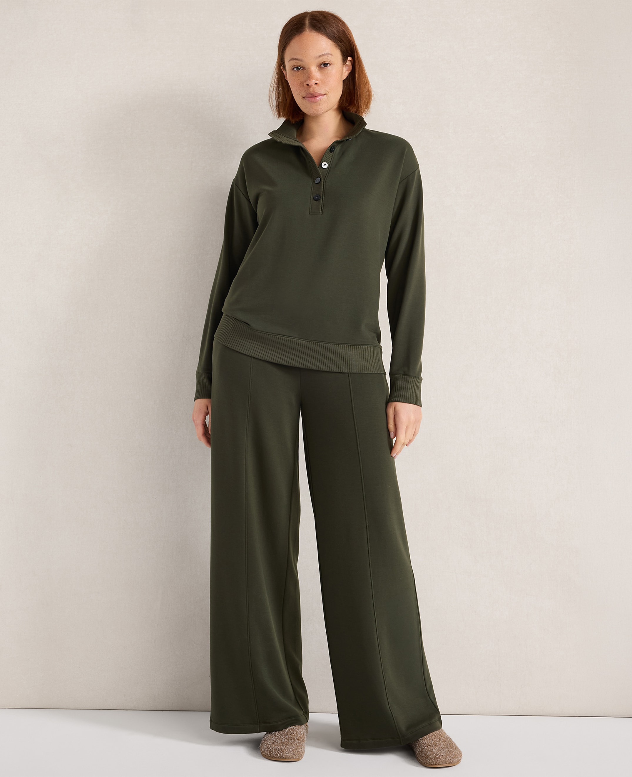 A woman stands in a dark green loungewear set with wide-leg pants and a collared top, against a plain, textured wall.