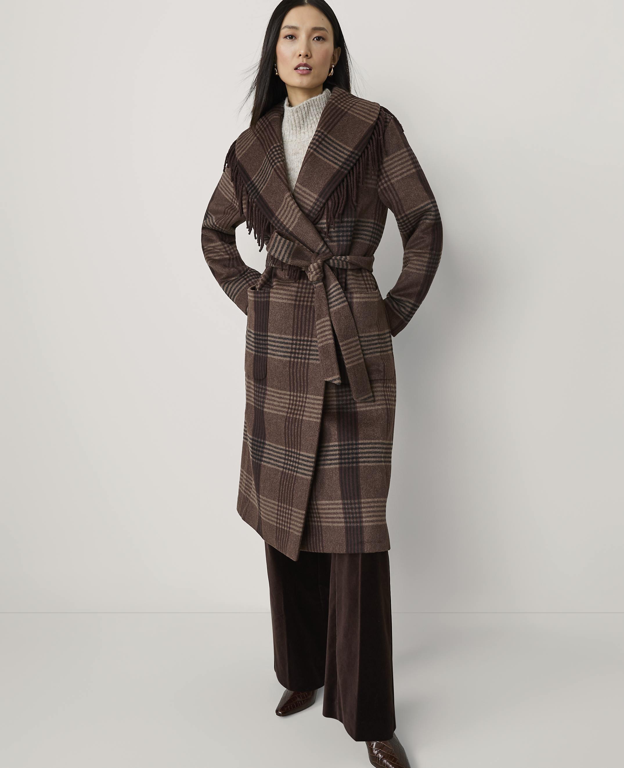 A woman in a brown plaid coat with fringe stands against a plain white background.
