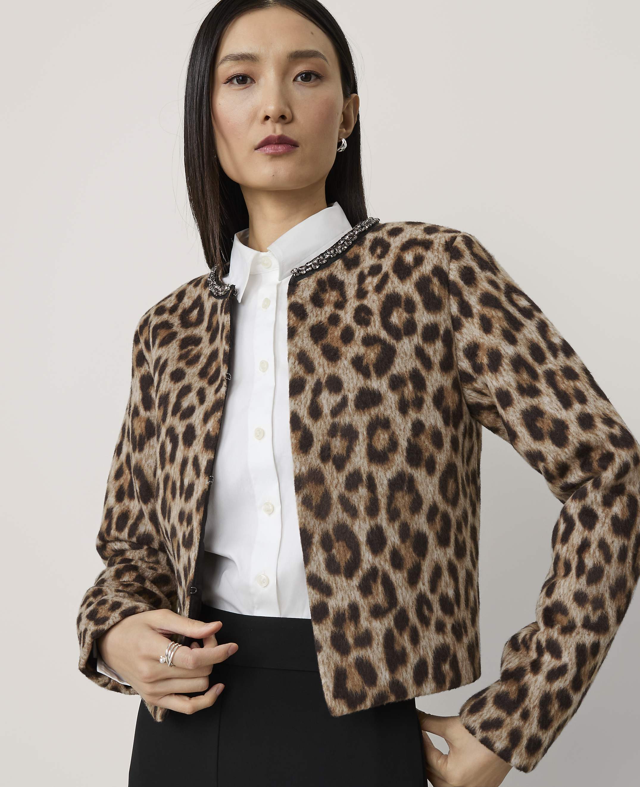 A woman in a leopard print jacket and white shirt stands against a plain, light gray background.