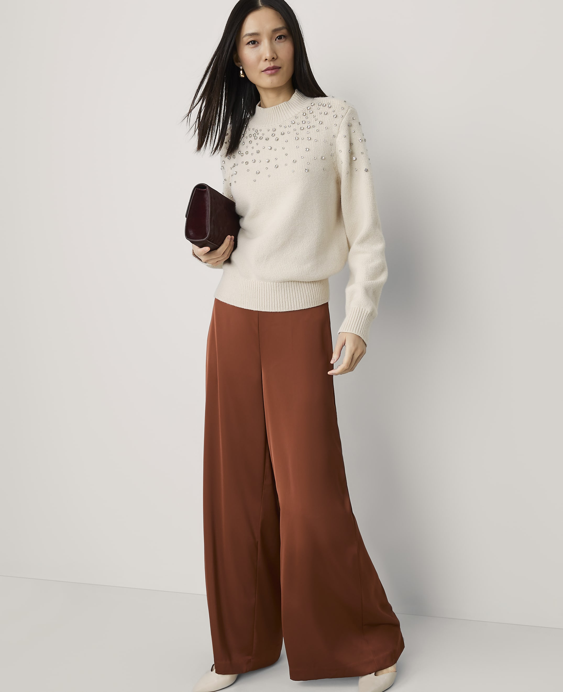 A woman in a cream embellished sweater and rust-colored wide-leg pants stands against a plain white background, holding a burgundy clutch.