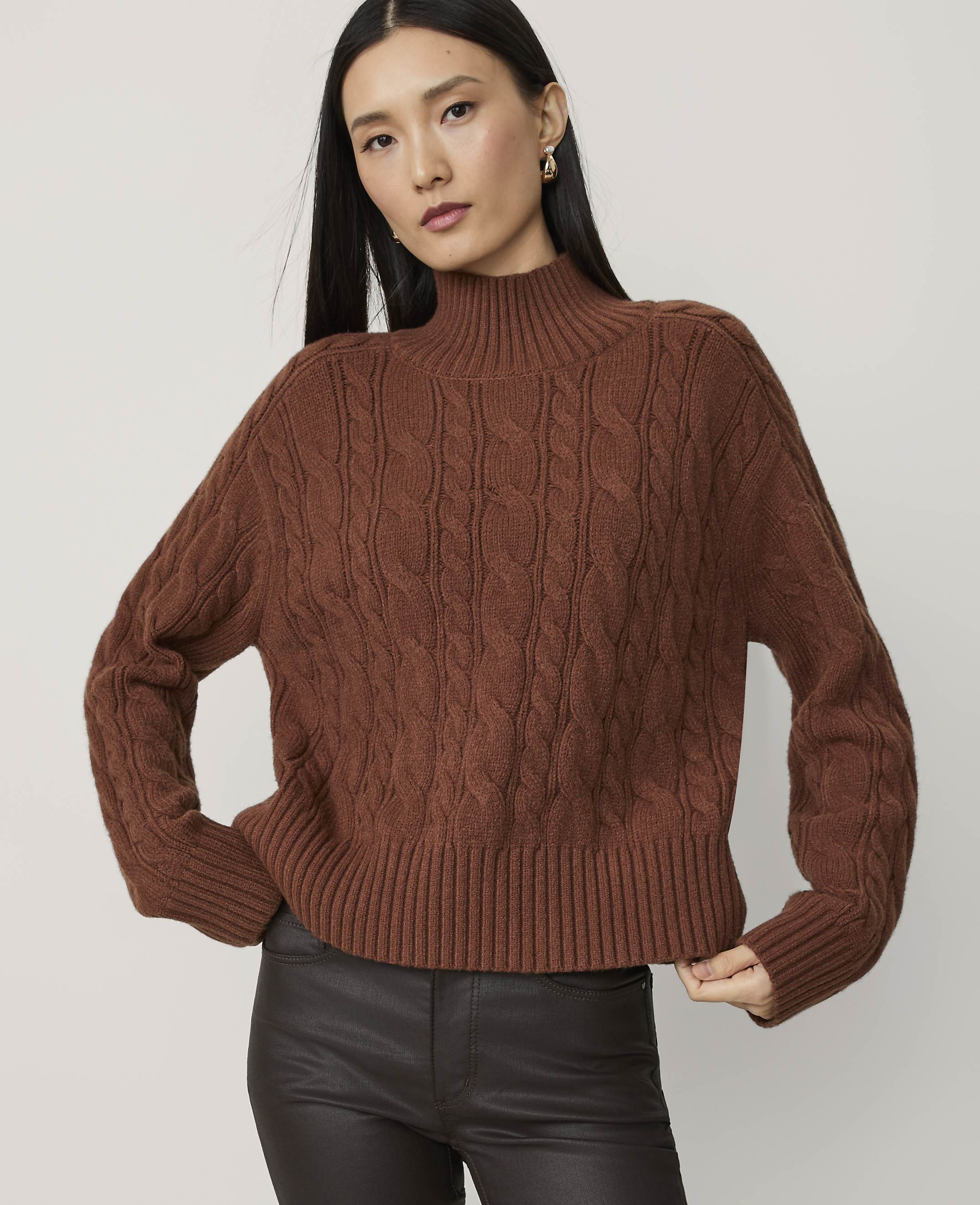 A woman in a brown cable-knit turtleneck sweater and dark pants poses against a plain white background.
