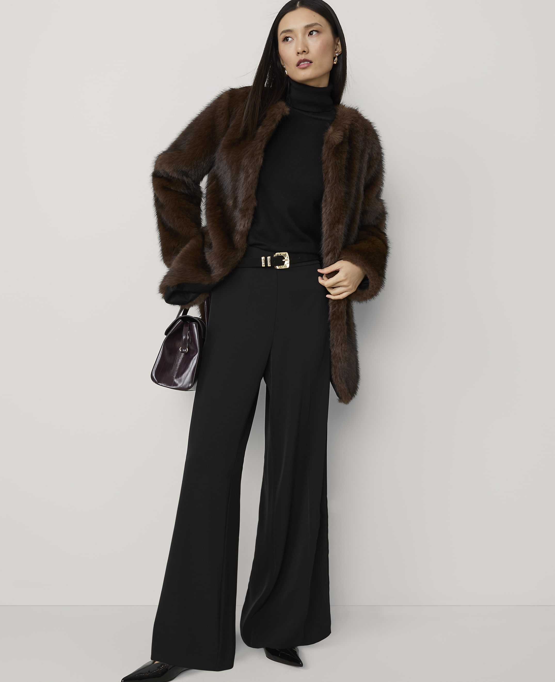 A woman in a brown fur coat, black turtleneck, and wide-leg pants stands against a plain white background, holding a dark handbag.