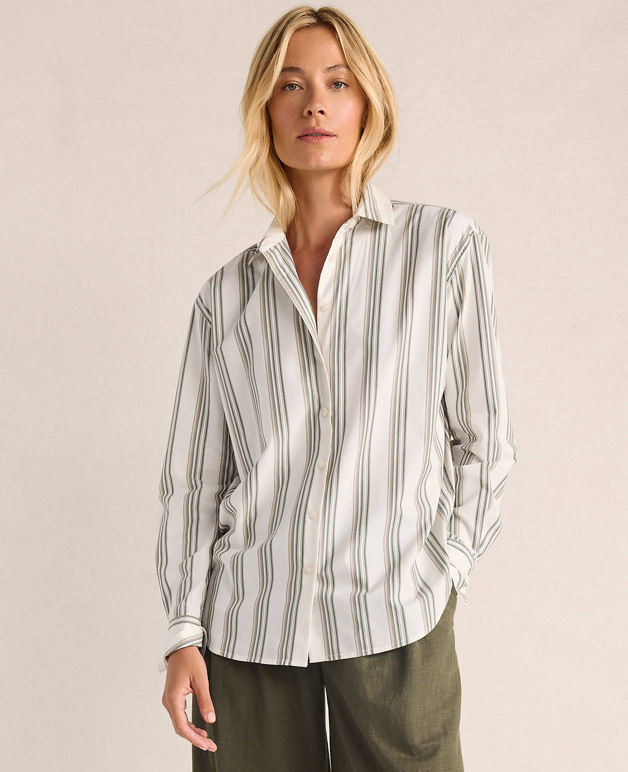A woman in a striped button-down shirt and green pants stands against a plain, light-colored background.