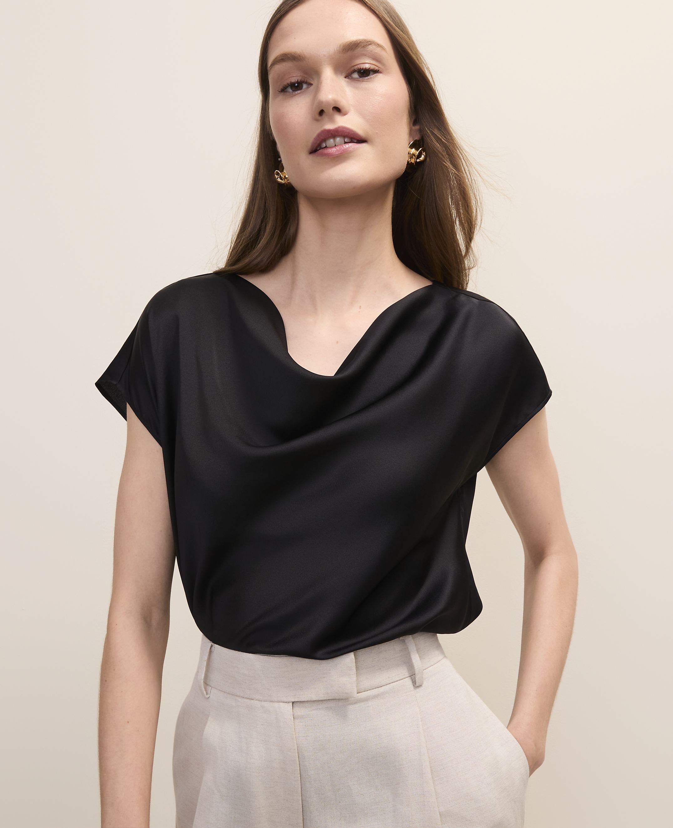 A woman in a black cowl-neck top and beige pants stands against a plain, light-colored background.