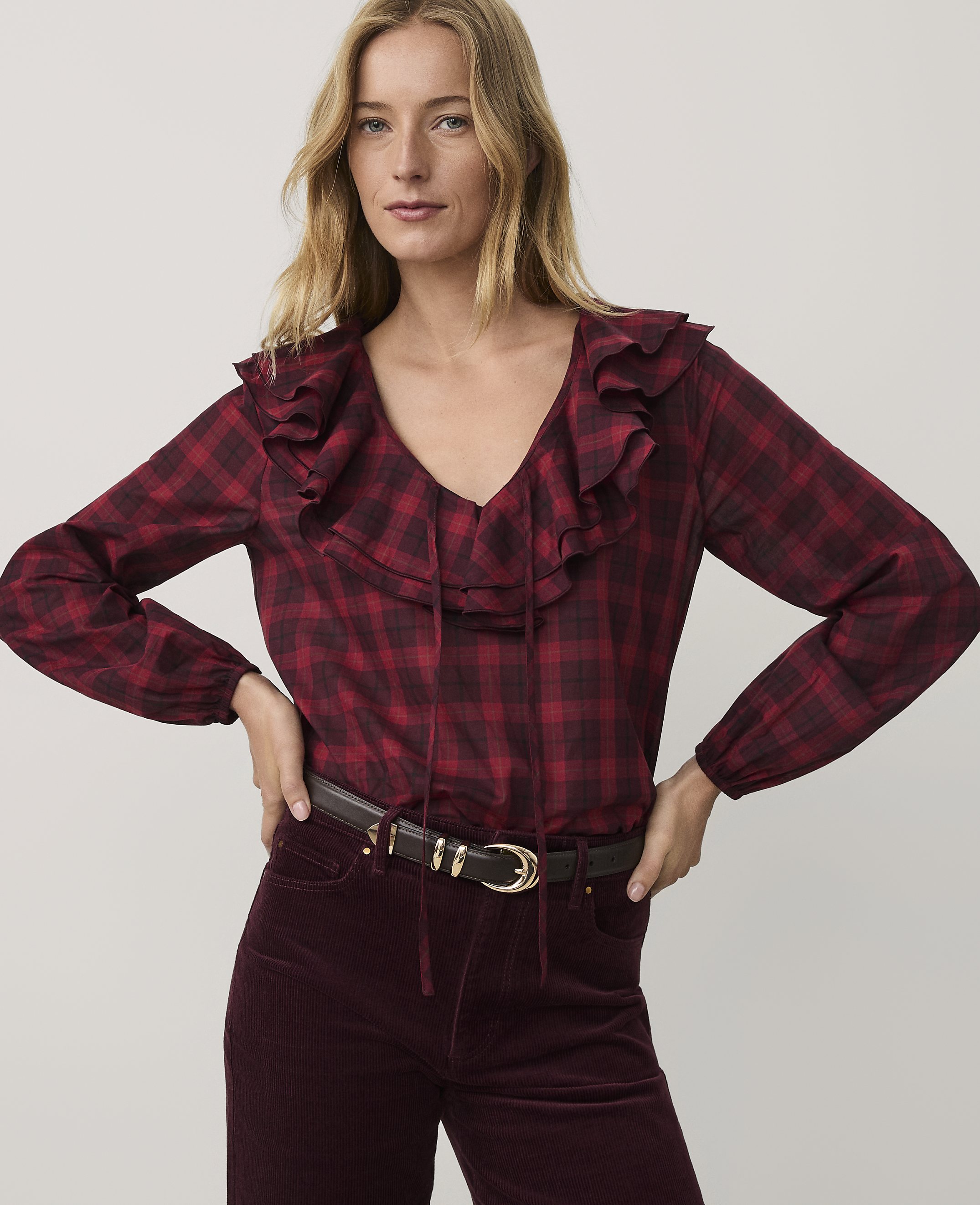 A woman wearing a red plaid ruffled blouse and burgundy corduroy pants stands against a plain light gray background.