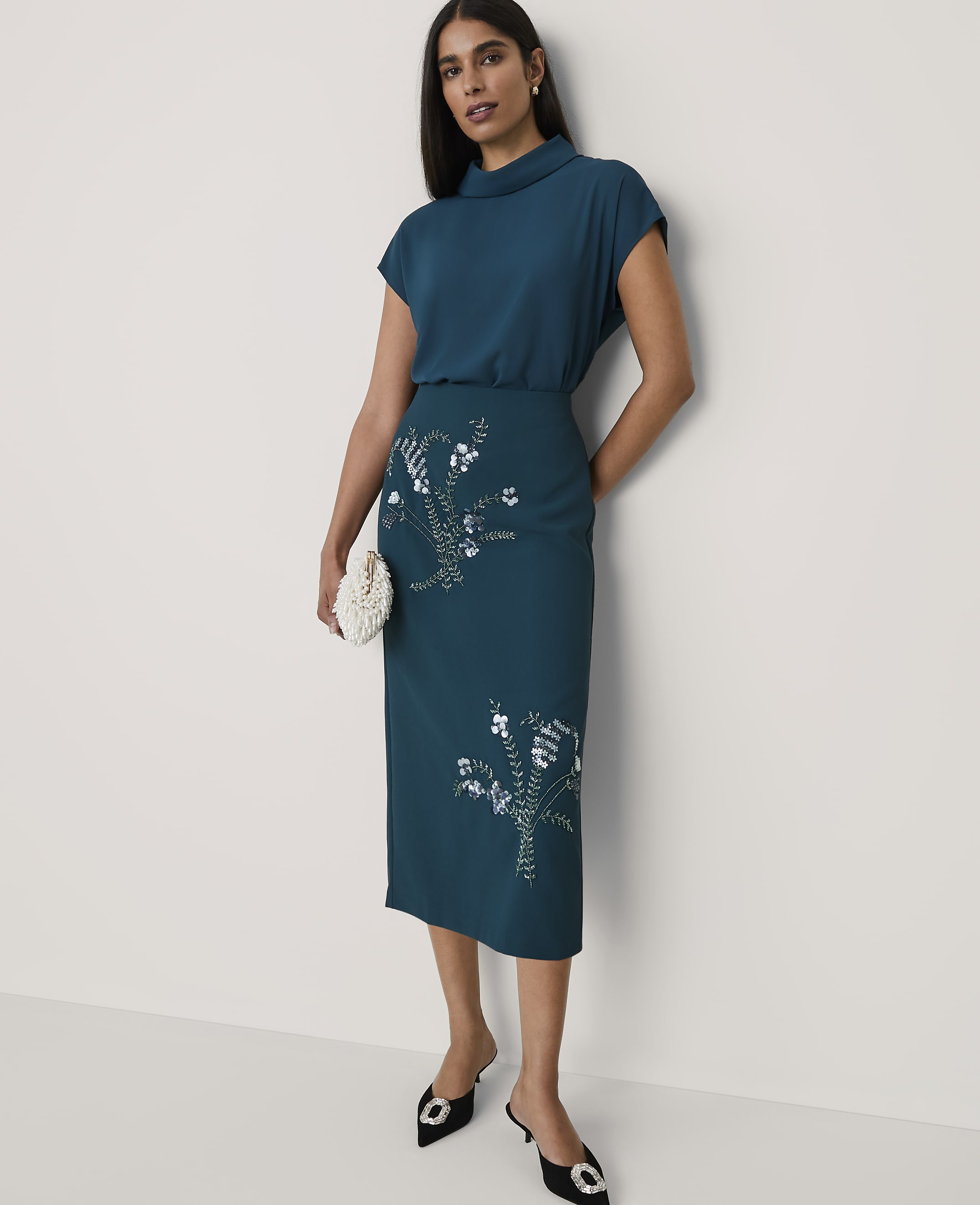A woman wearing a teal two-piece outfit with floral sequins and black heels stands against a plain white background.