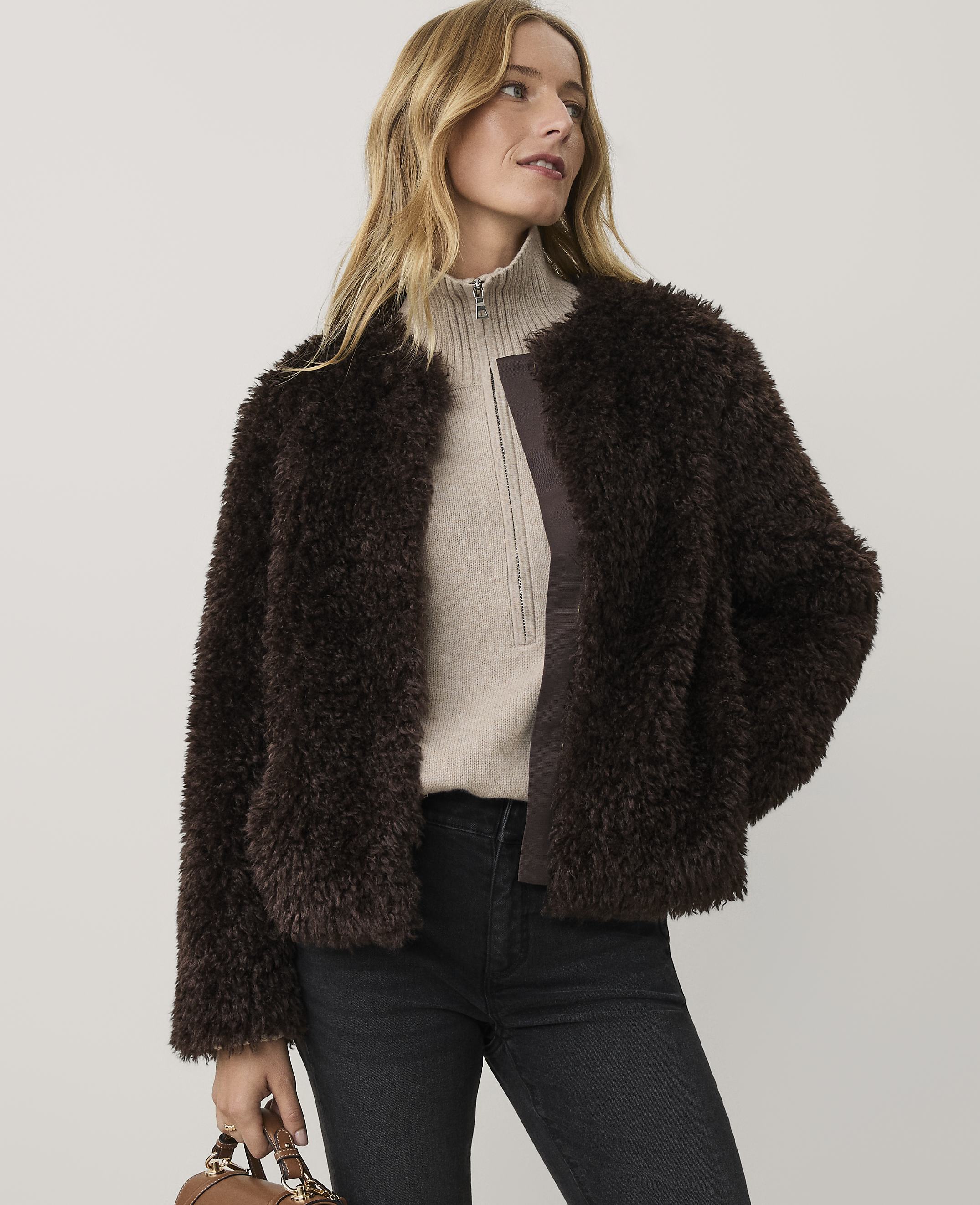A woman wearing a dark brown faux-fur jacket, a beige quarter-zip sweater, and black jeans stands against a plain light gray background.