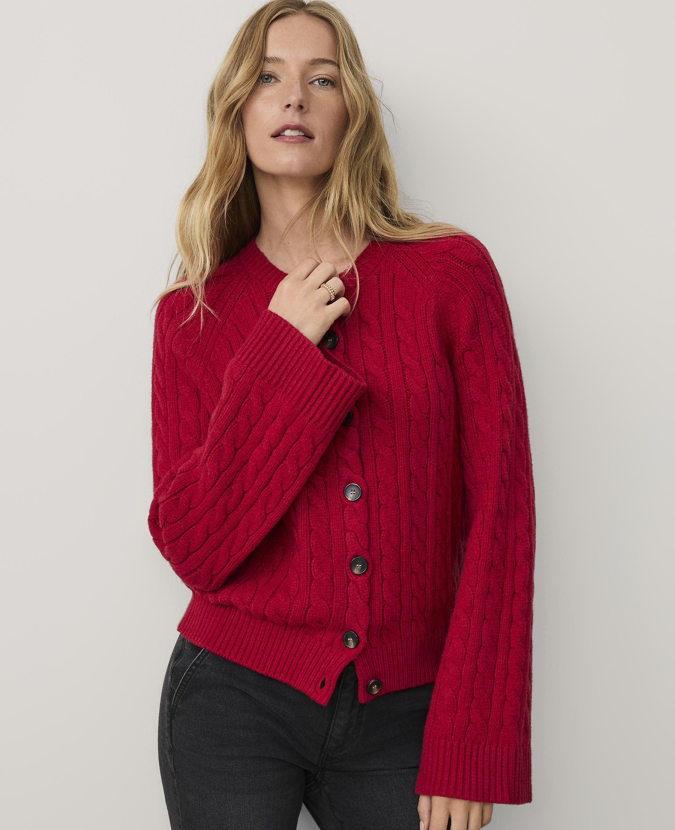 A woman wearing a red cable-knit cardigan and black jeans stands against a plain, light-colored background.