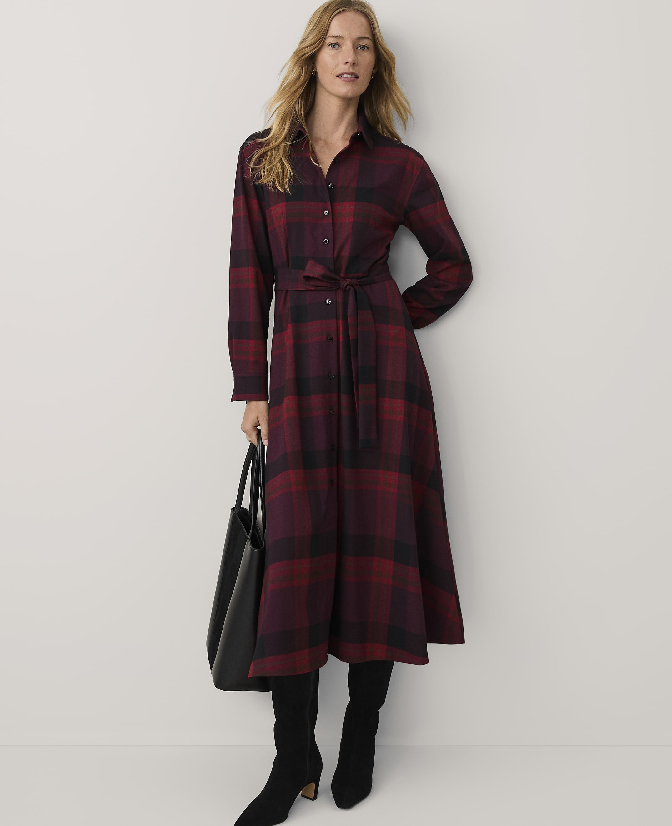 A woman wearing a red and black plaid maxi dress and black boots stands against a plain white background while holding a black handbag.