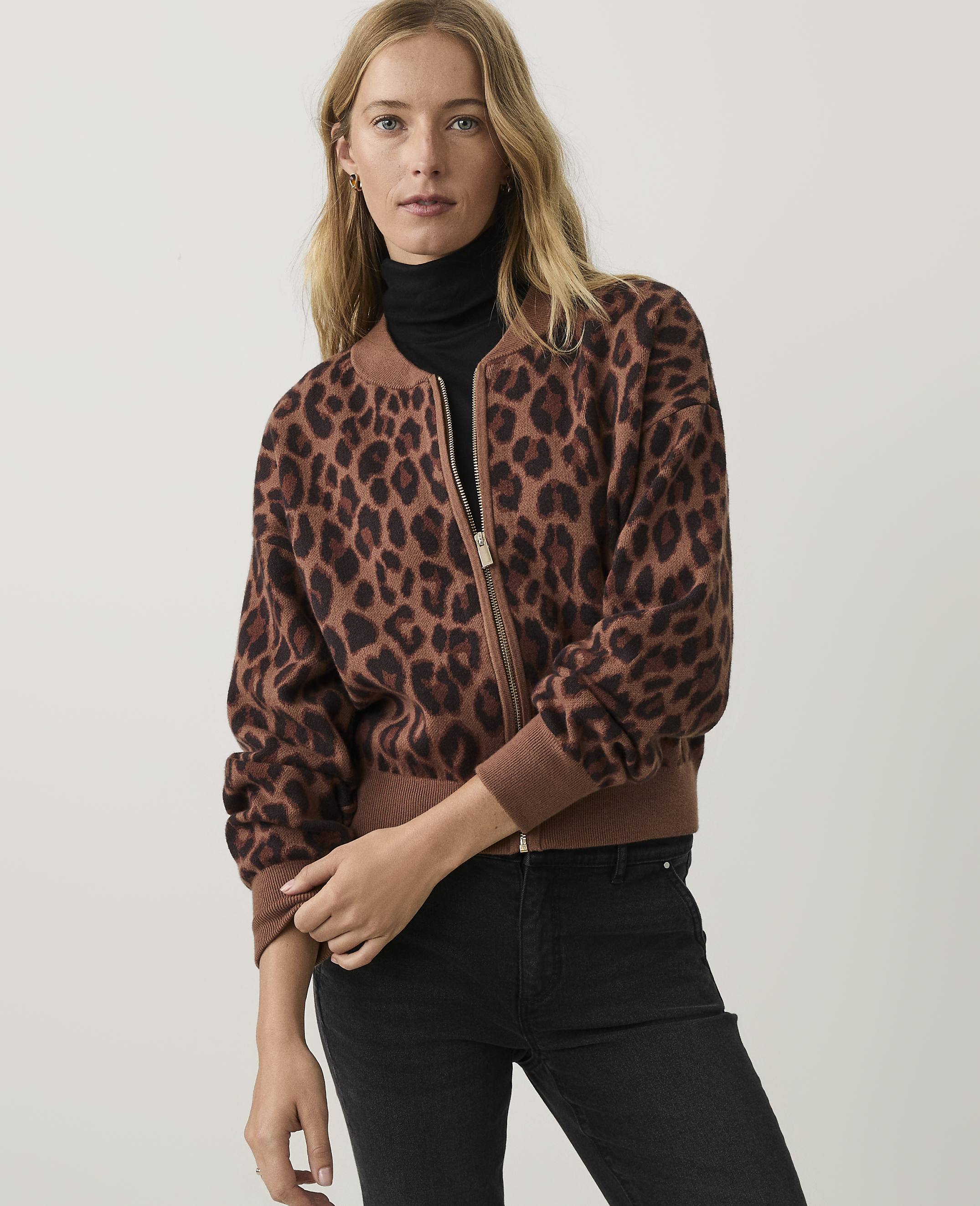 A woman wearing a brown leopard-print bomber jacket, black turtleneck, and black jeans stands against a plain light-grey background.