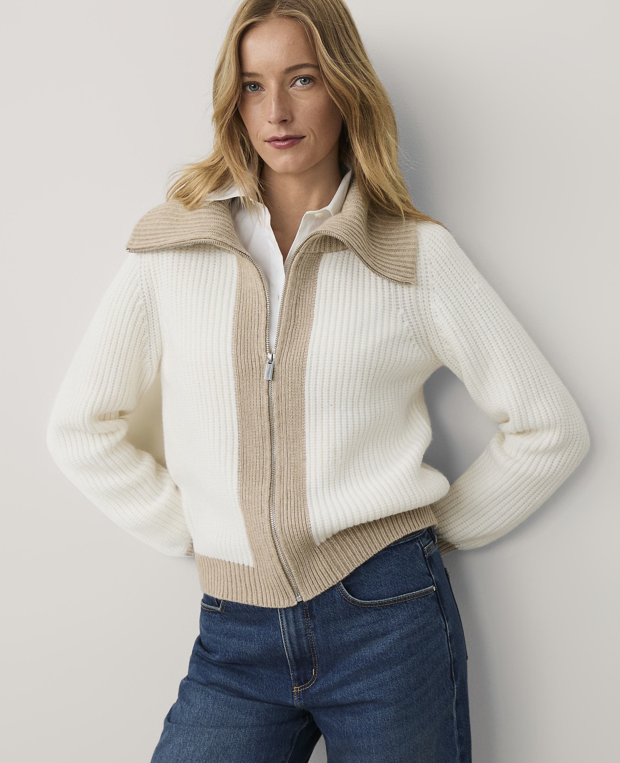 A woman wearing a white and beige ribbed zip-up sweater and blue jeans stands against a plain light gray wall.