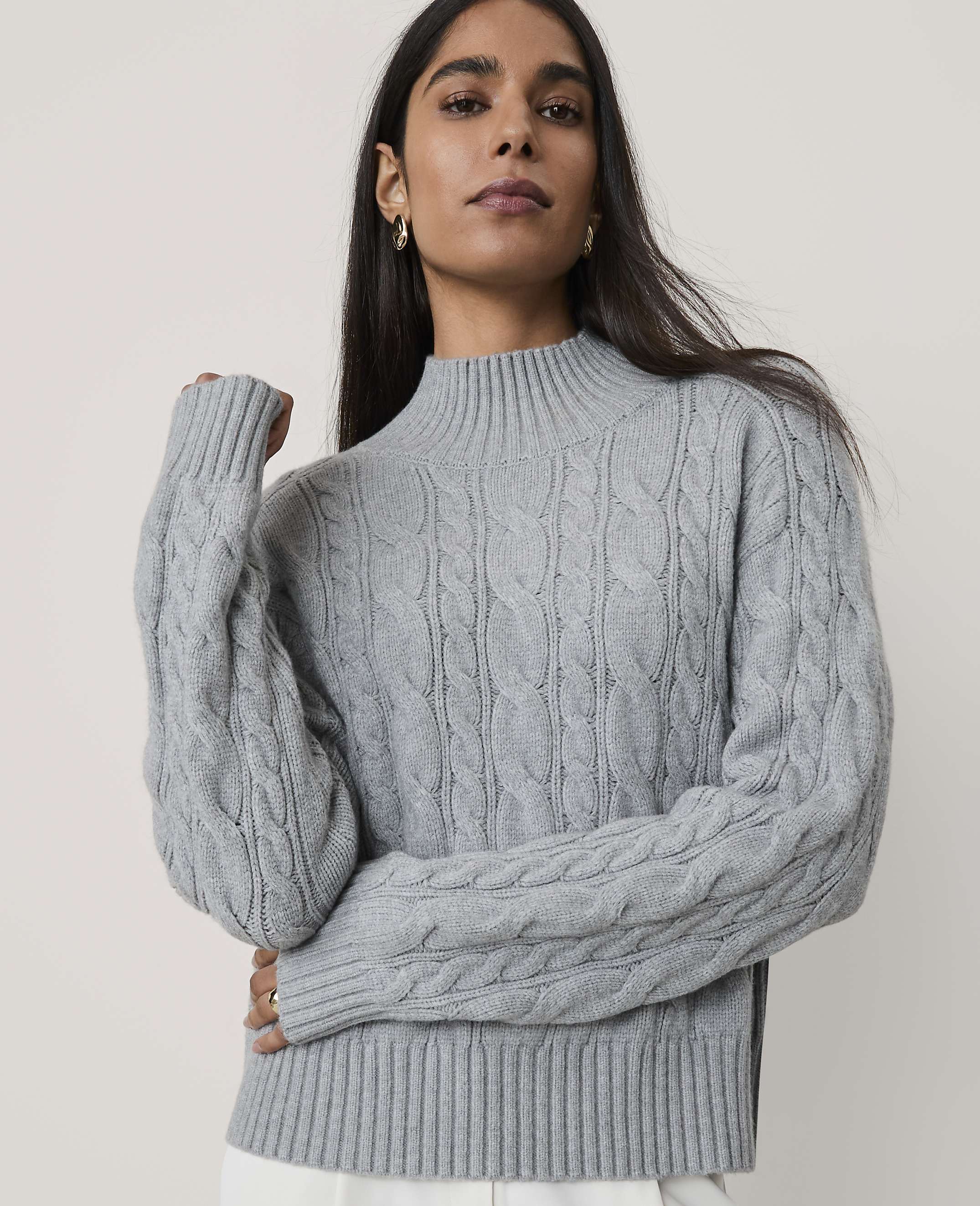 A woman wearing a grey cable-knit sweater stands against a plain, light-colored background.
