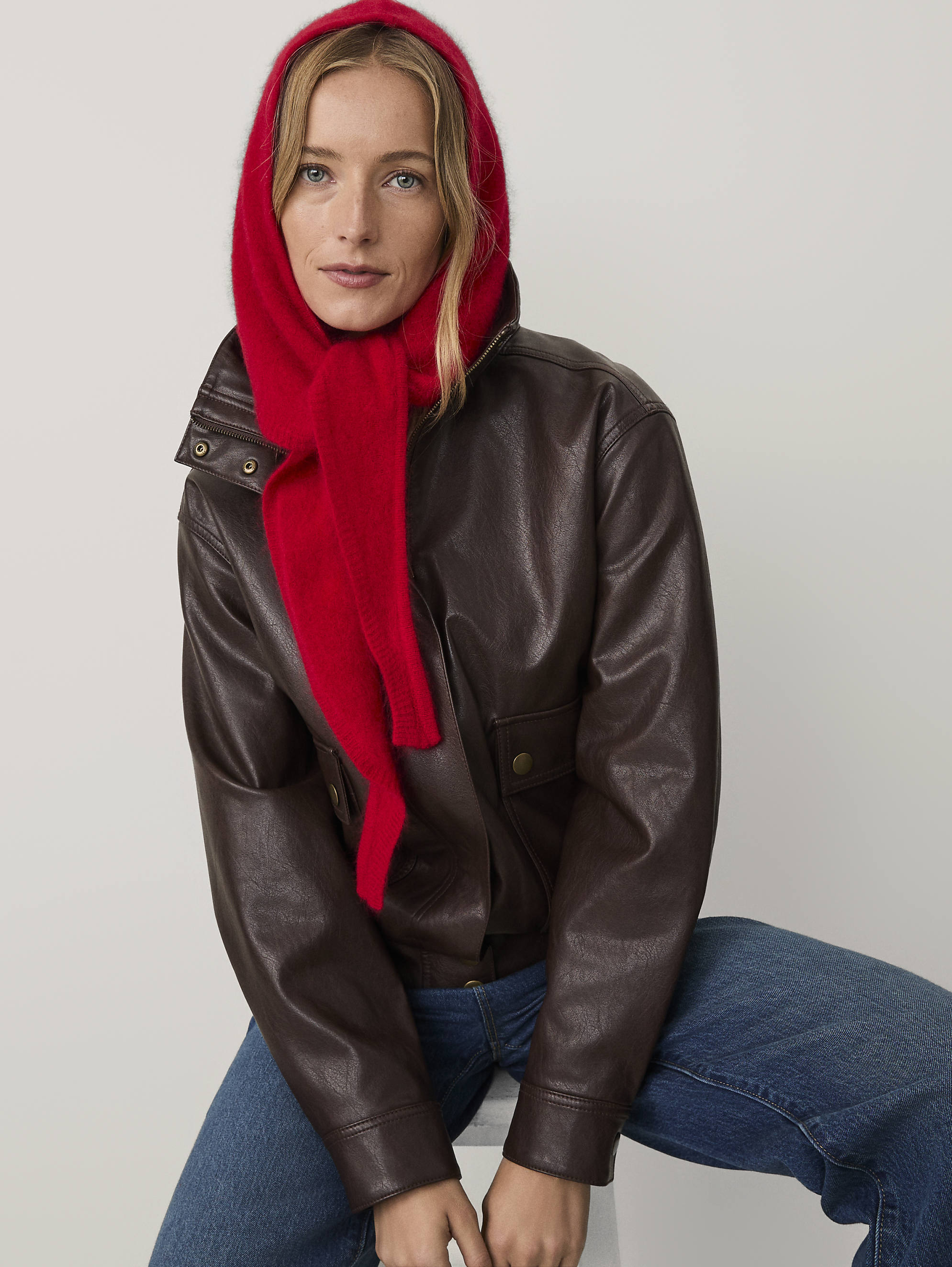 A woman wearing a red hood, a brown leather jacket, and blue jeans poses against a plain light gray background.