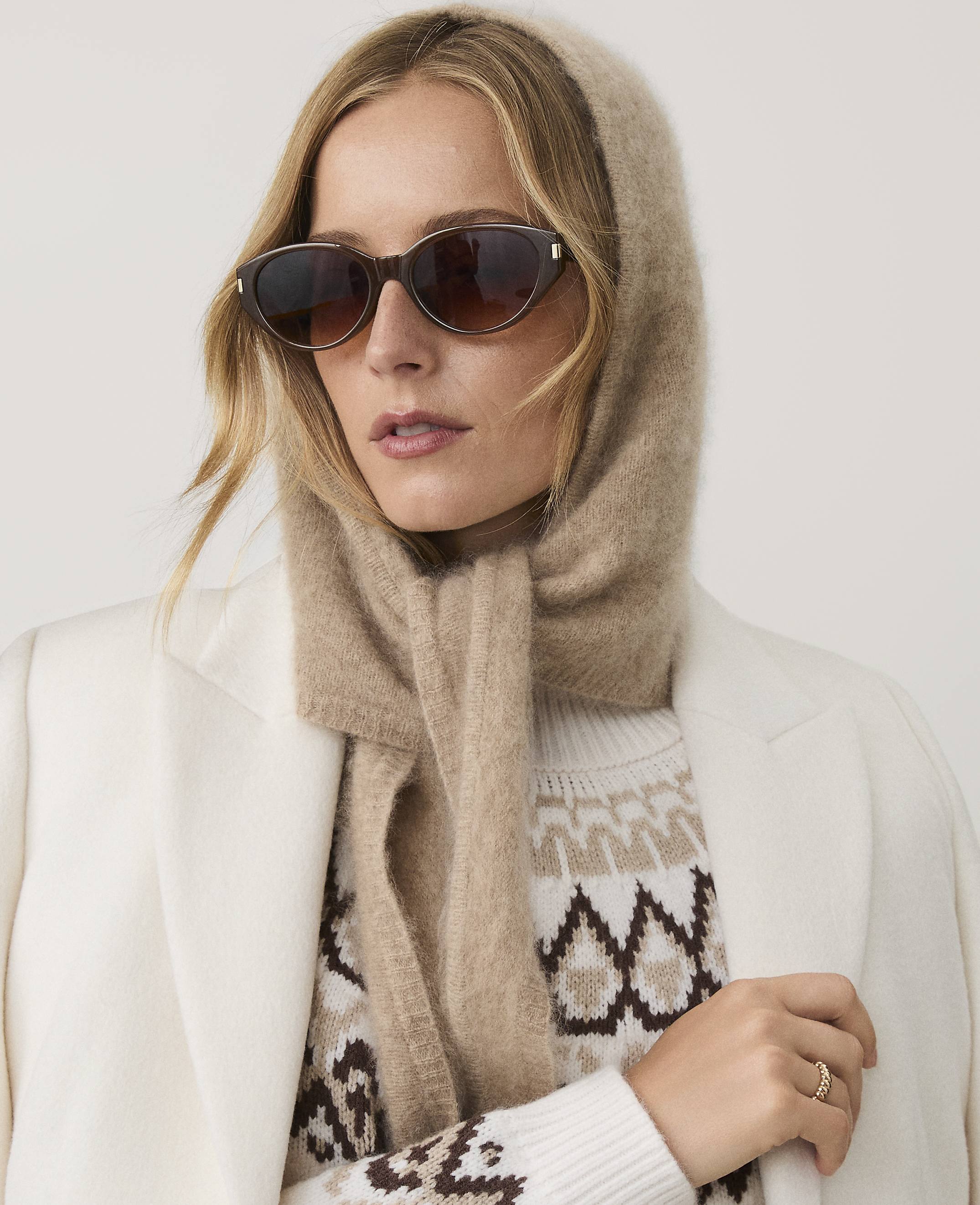 A woman wearing sunglasses, a beige hood-scarf, and a patterned sweater under a white coat poses against a plain light background.