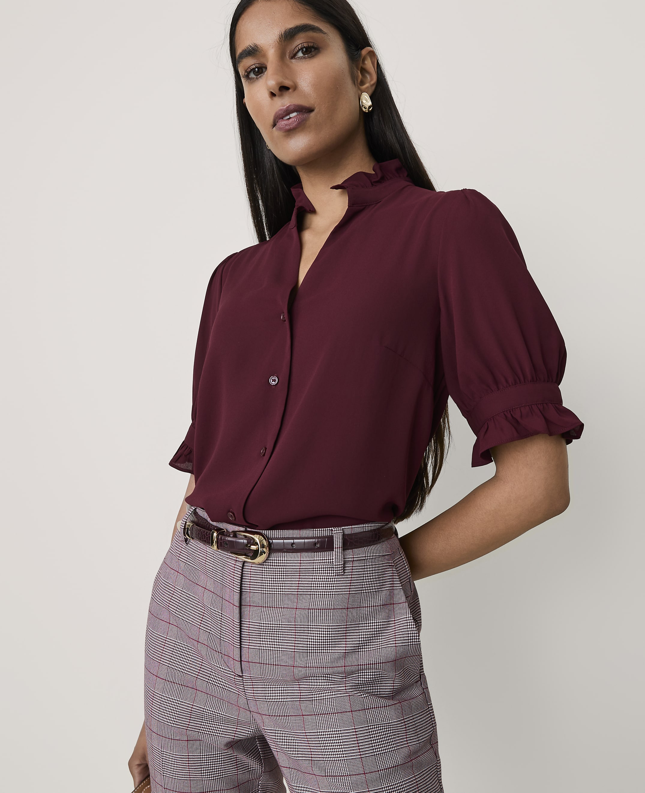 A woman wearing a burgundy blouse and plaid trousers stands against a plain, light-colored background.