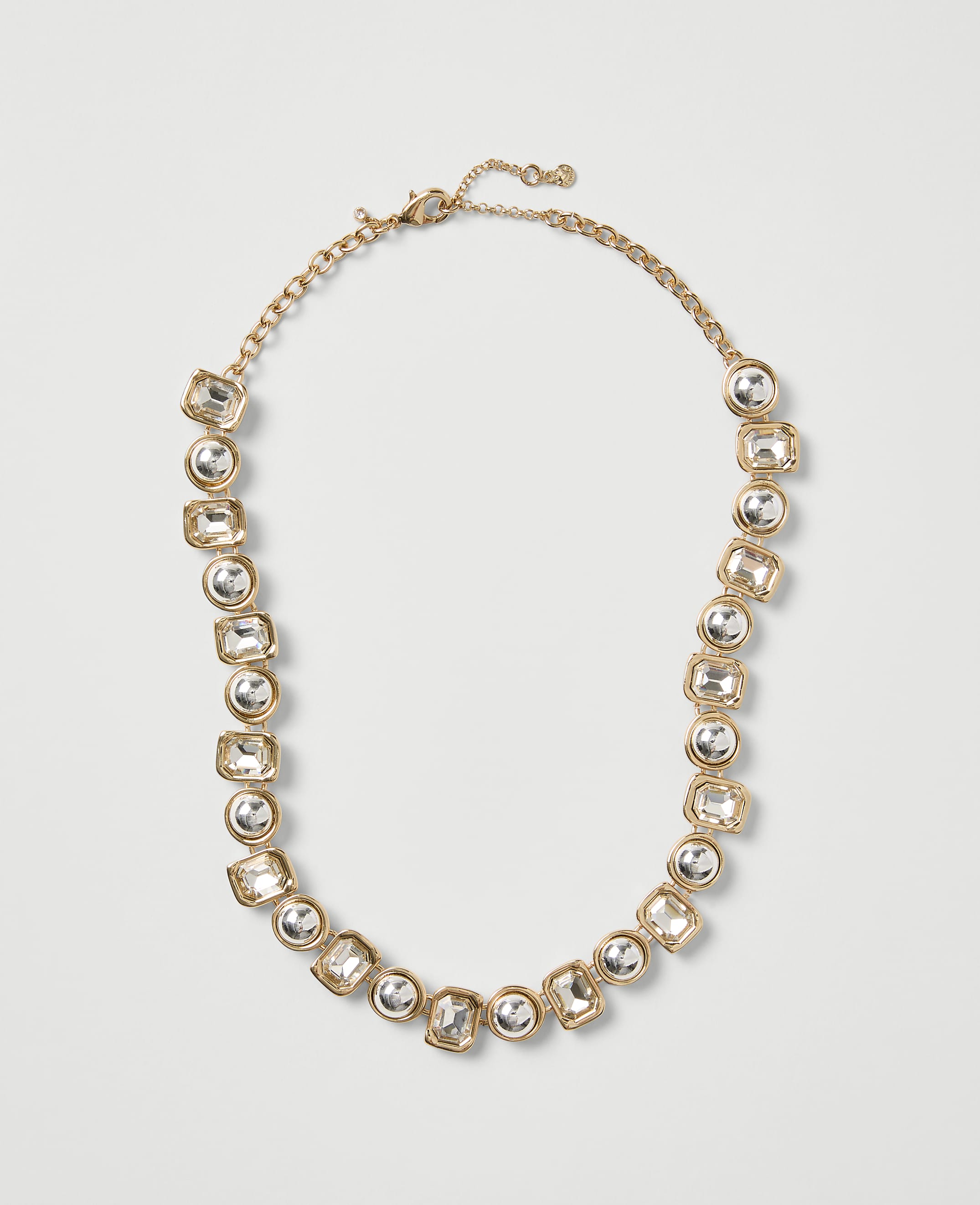 A gold-toned necklace with clear crystal stones is centered against a plain light gray background.
