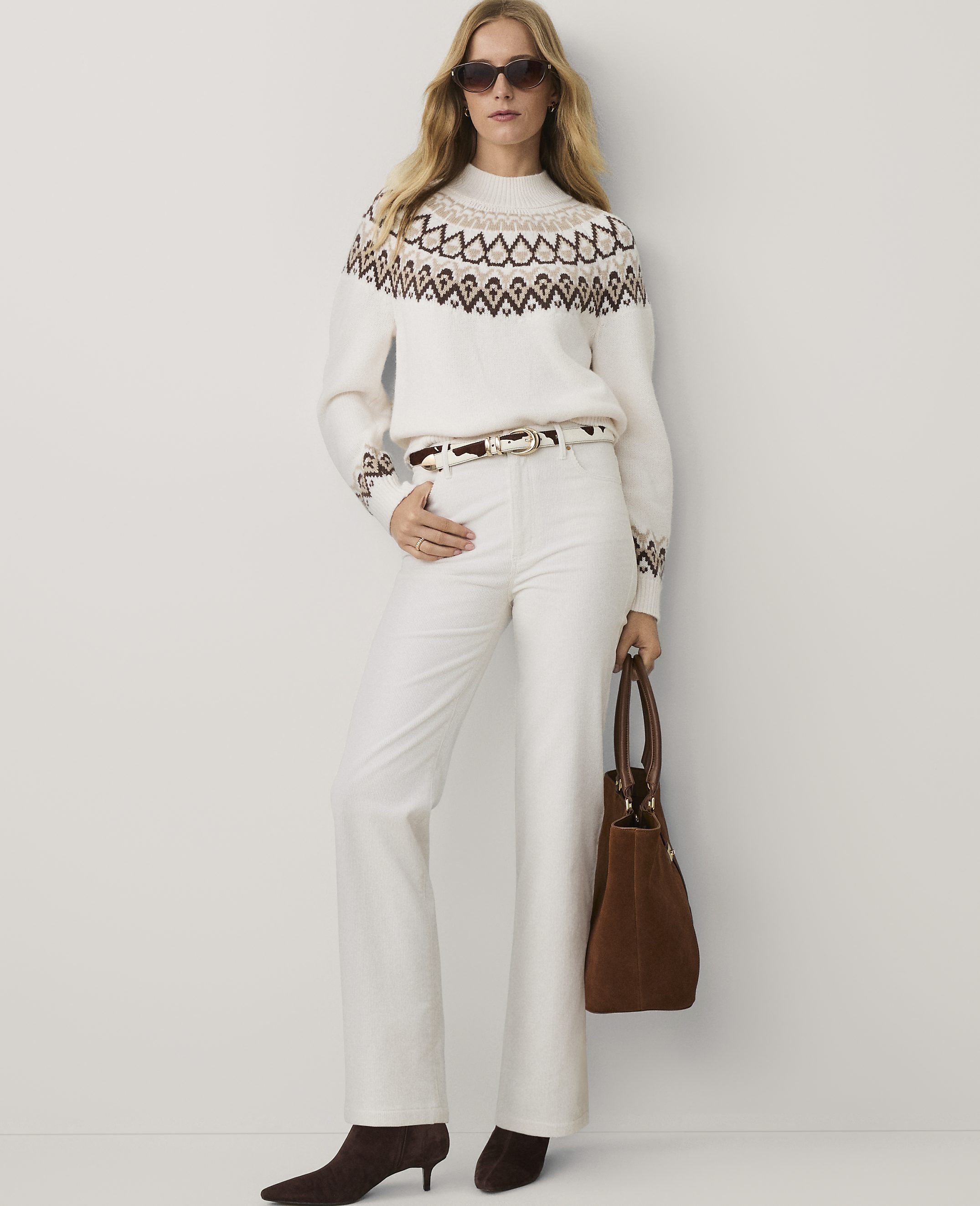 A woman wearing a patterned white sweater, white trousers, and brown heels stands against a plain white background.