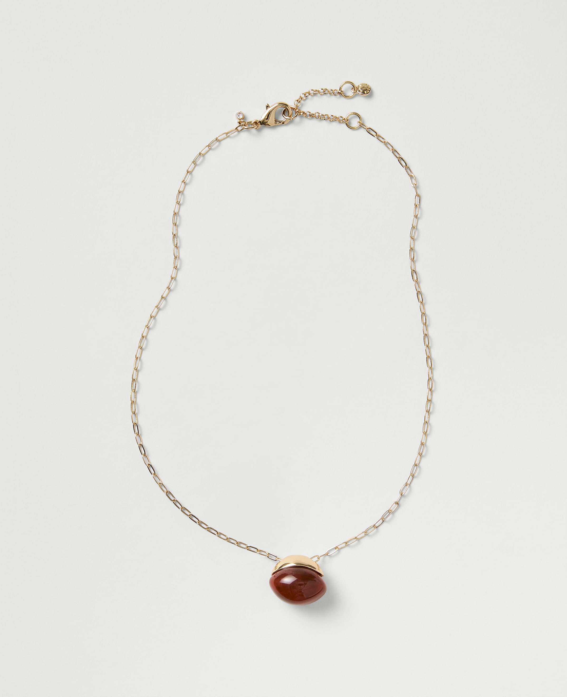 A gold chain necklace with a red oval gemstone pendant rests against a plain light gray background.