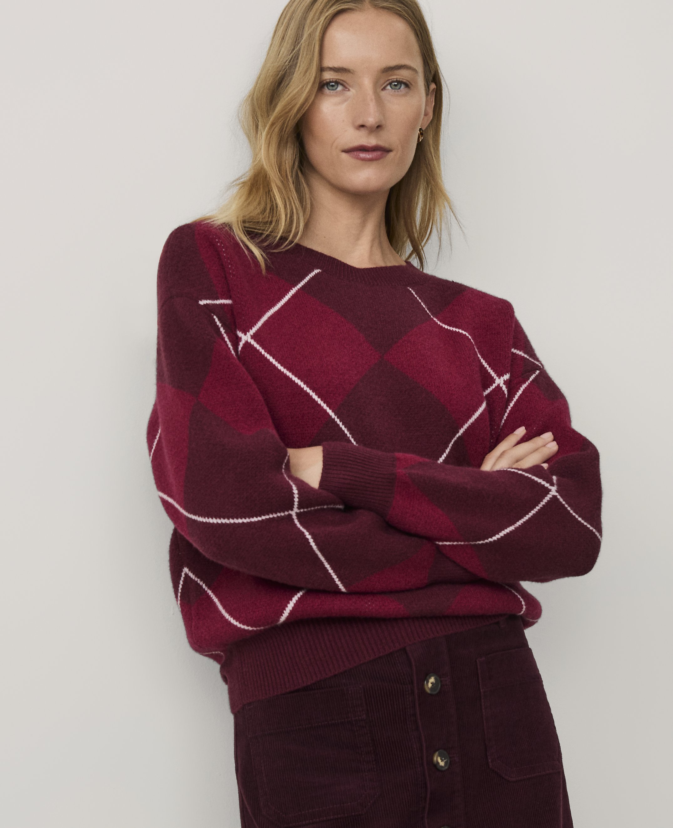 A blonde woman wearing a red and burgundy argyle sweater and matching corduroy skirt stands with crossed arms against a plain white background.