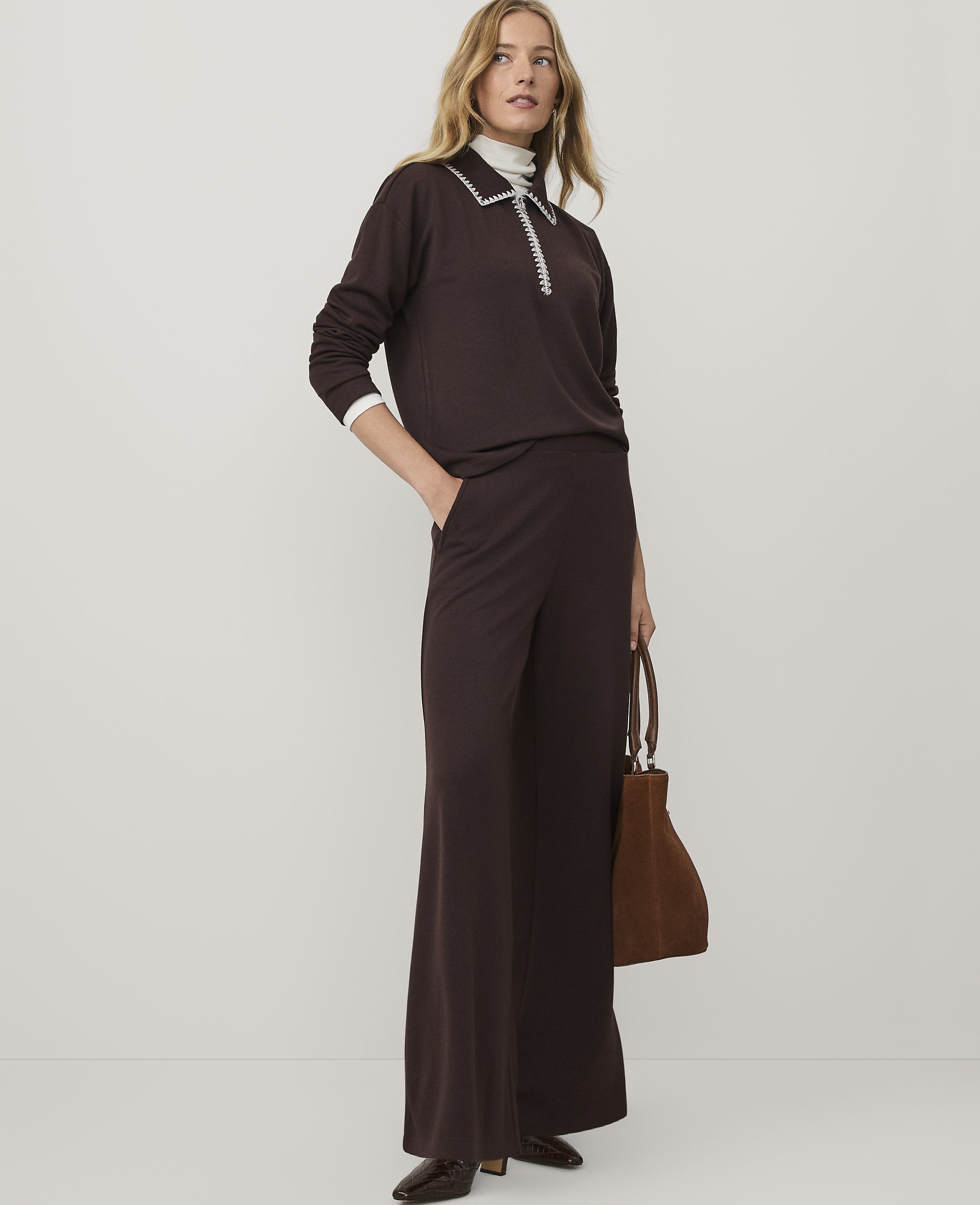 A woman wearing a dark brown two-piece outfit and holding a brown handbag stands against a plain white background.