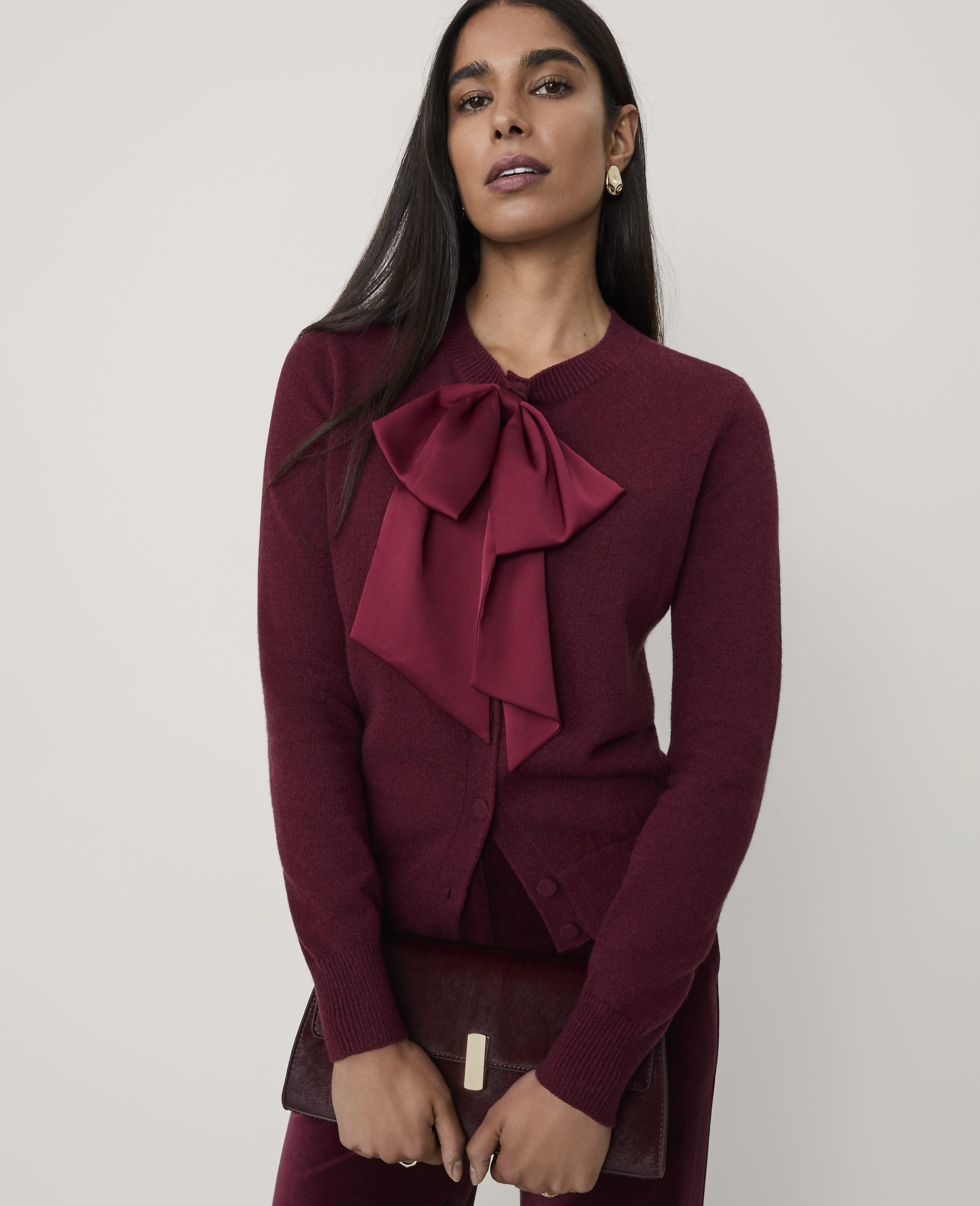 A woman wearing a burgundy cardigan with a large bow and matching trousers stands against a plain, light-colored background.