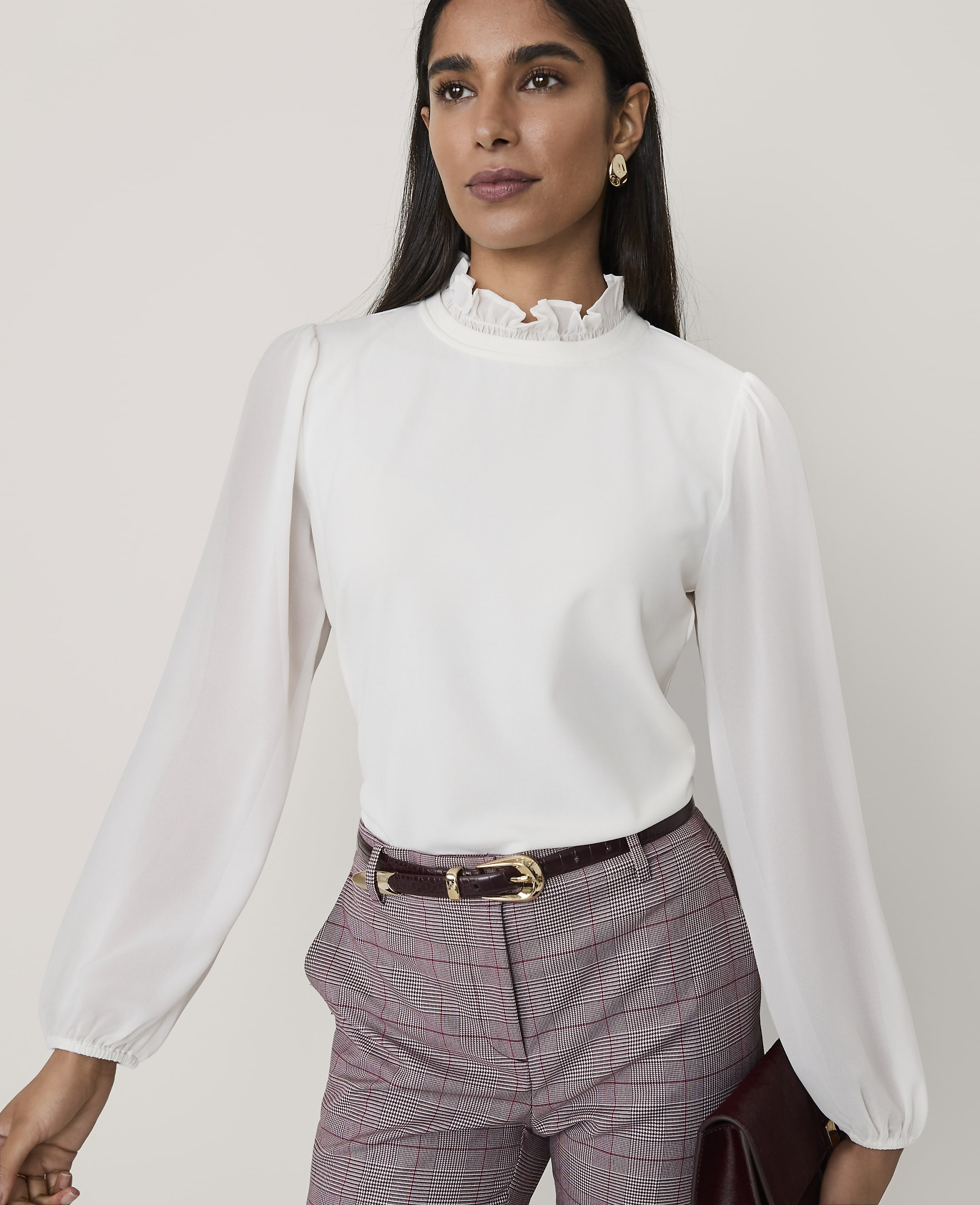 A woman wearing a white ruffled blouse and plaid trousers stands against a plain, light-colored background.