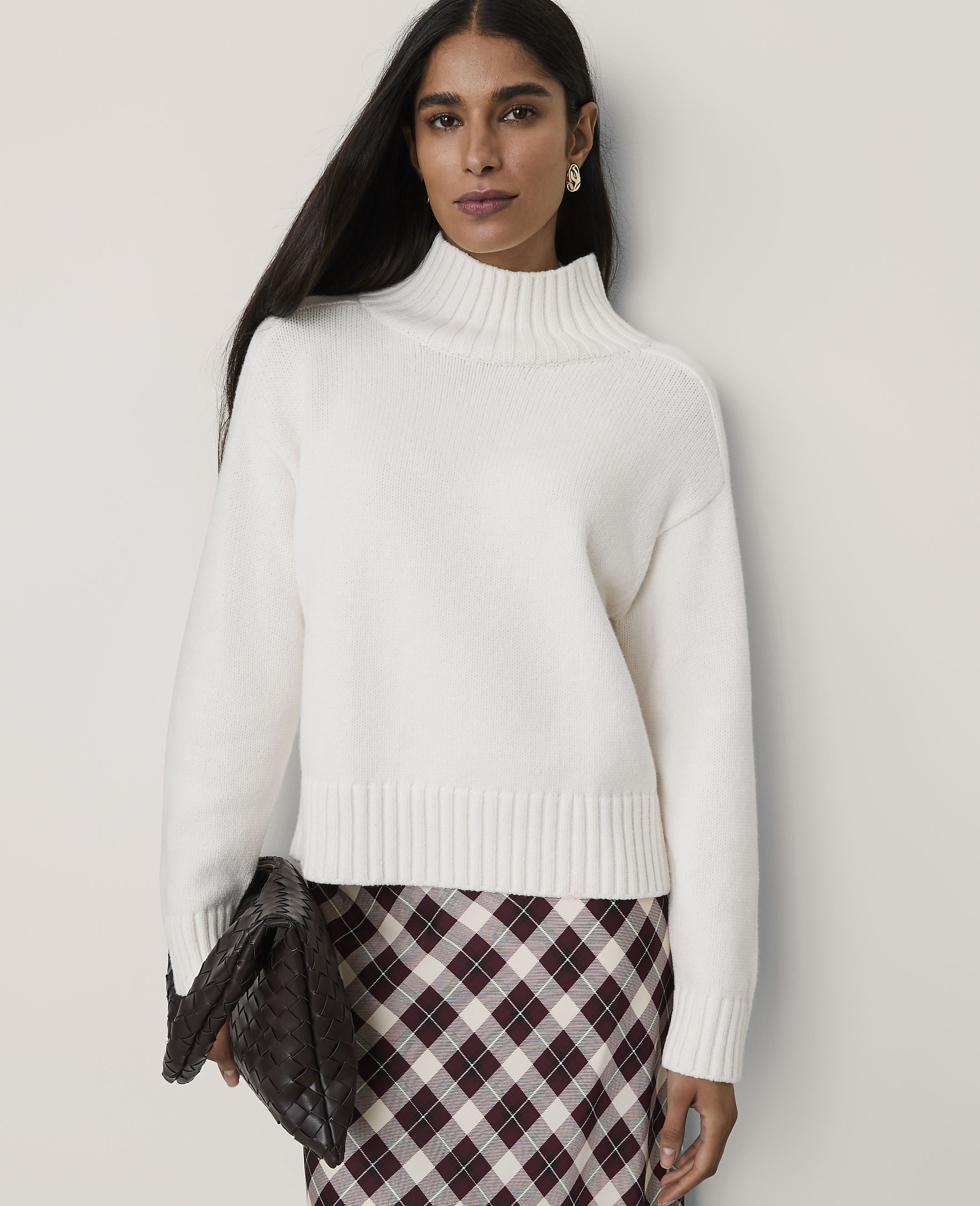 A woman wearing a white mock-neck sweater and a plaid skirt stands against a plain, light-colored wall.