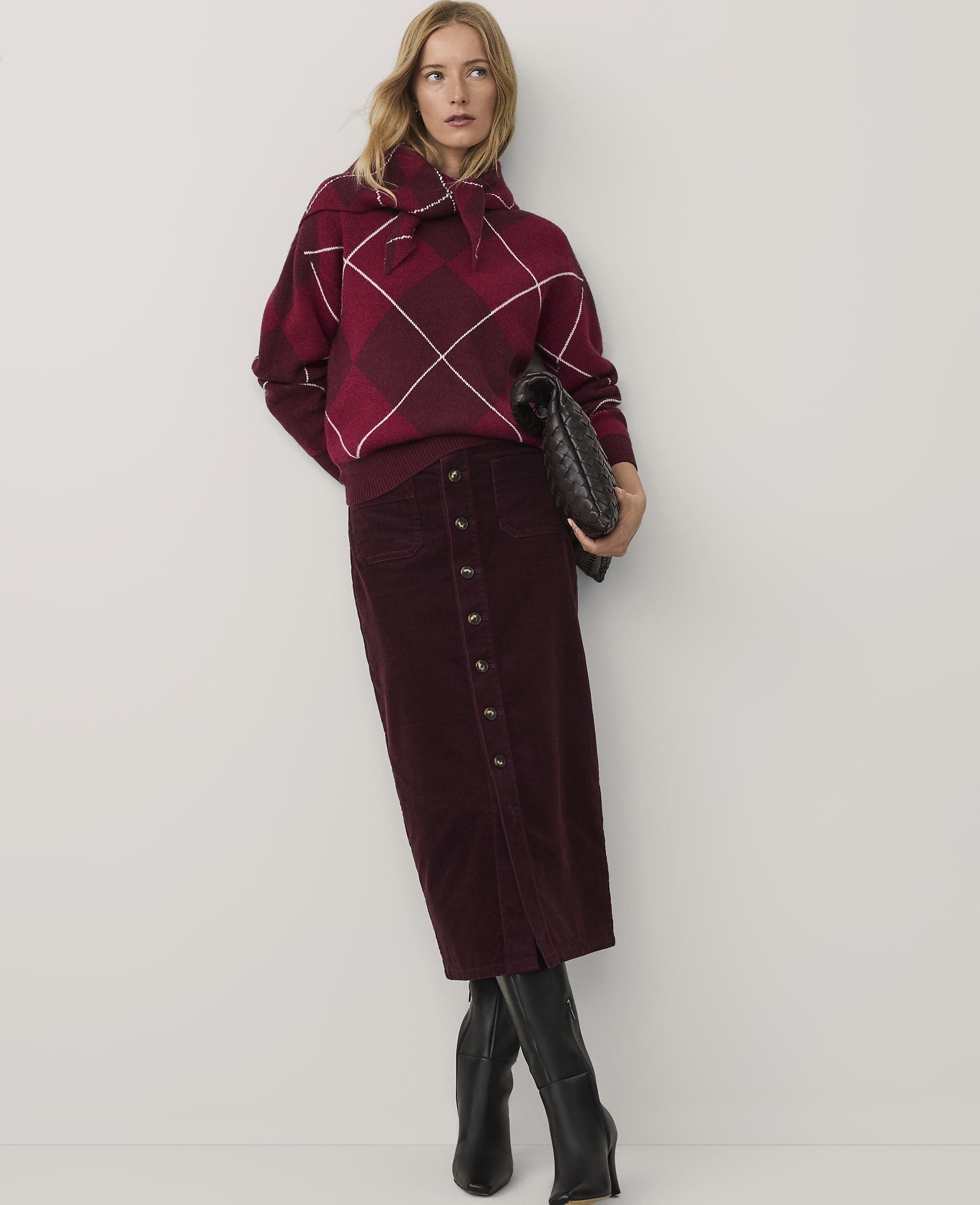 A woman wearing a red plaid sweater and a long burgundy skirt stands against a plain white background.