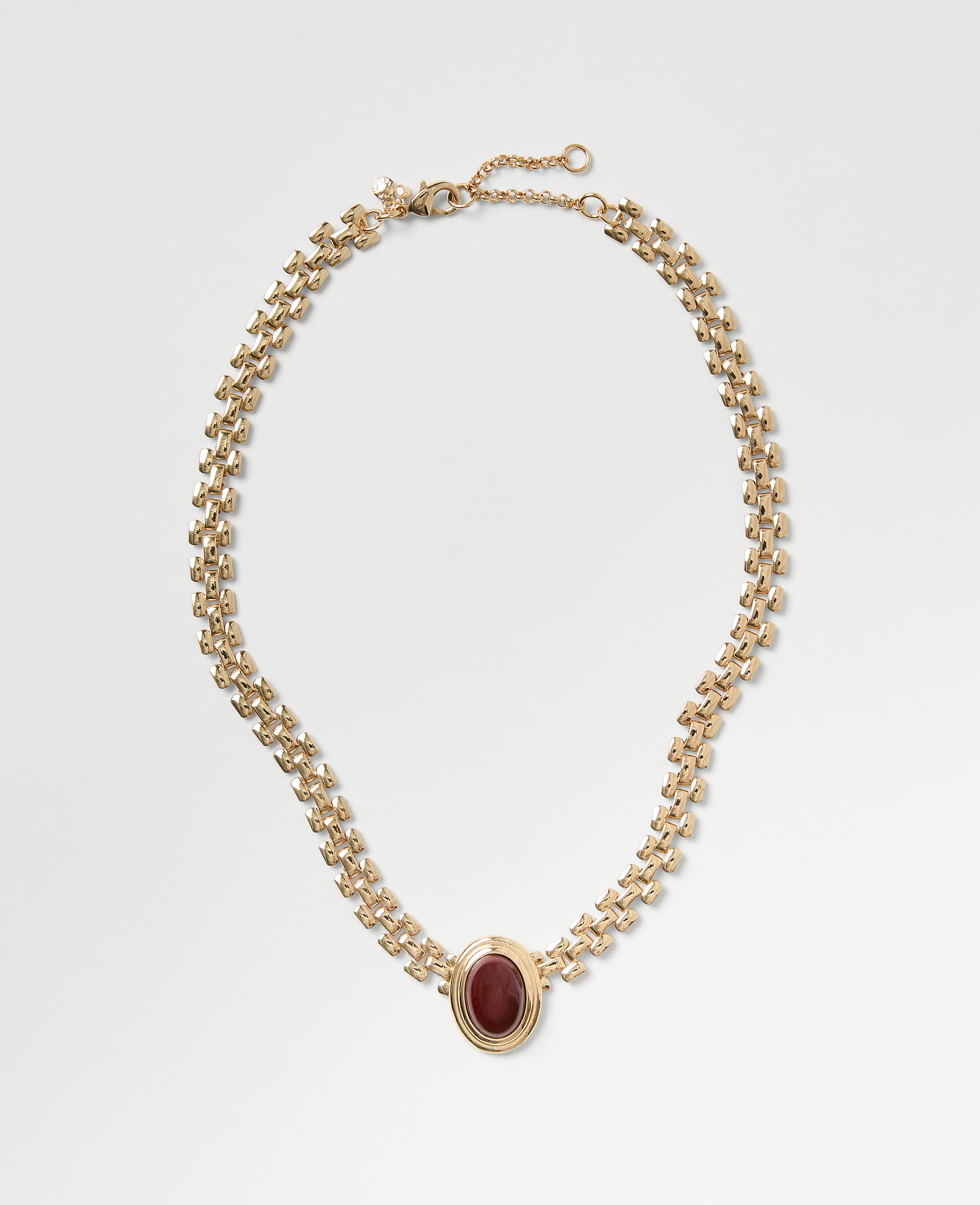 A gold chain necklace with a red oval gemstone pendant rests on a plain light gray background.